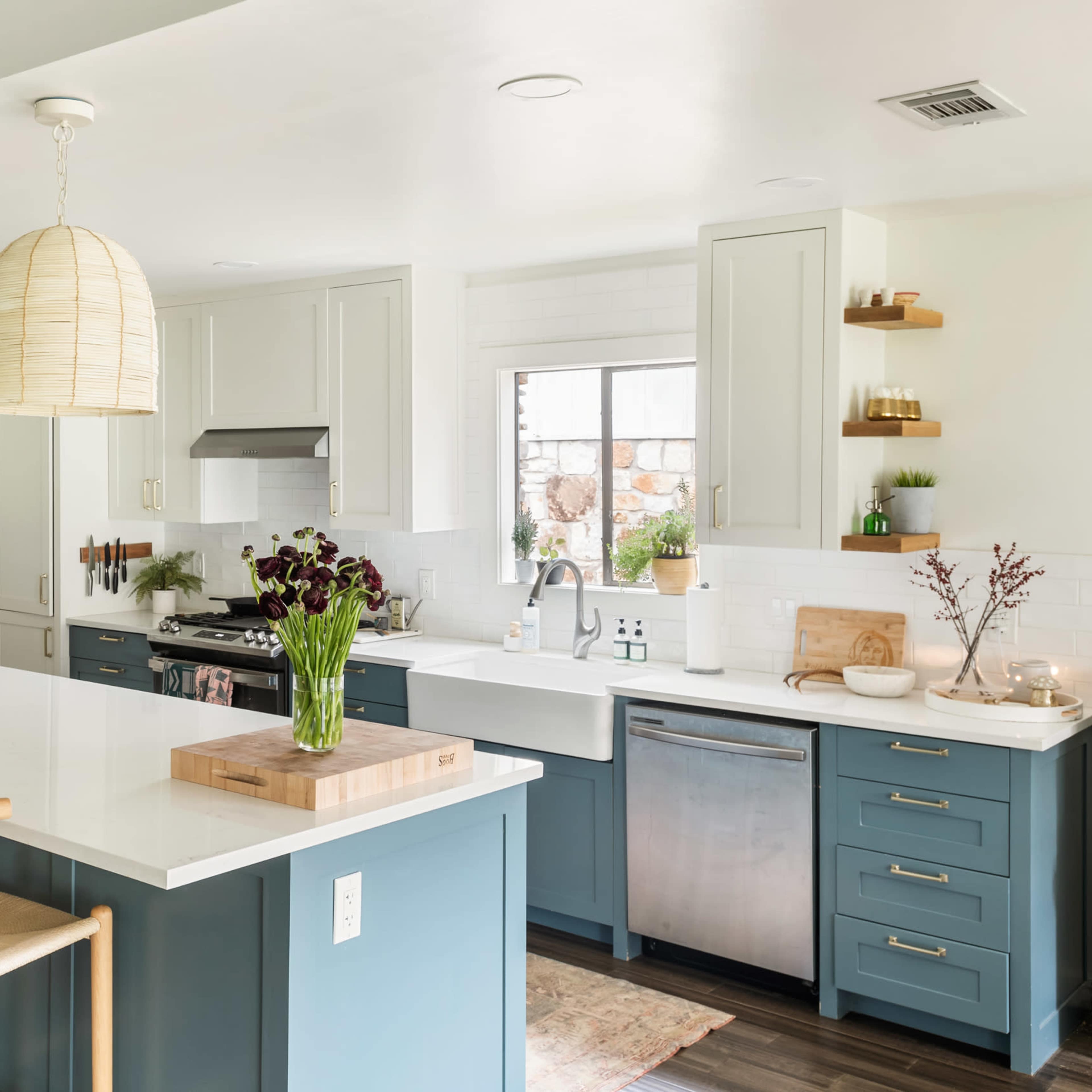 A modern kitchen features a central island with bar stools, white cabinetry, a stainless steel dishwasher, and decorative open shelving with plants.