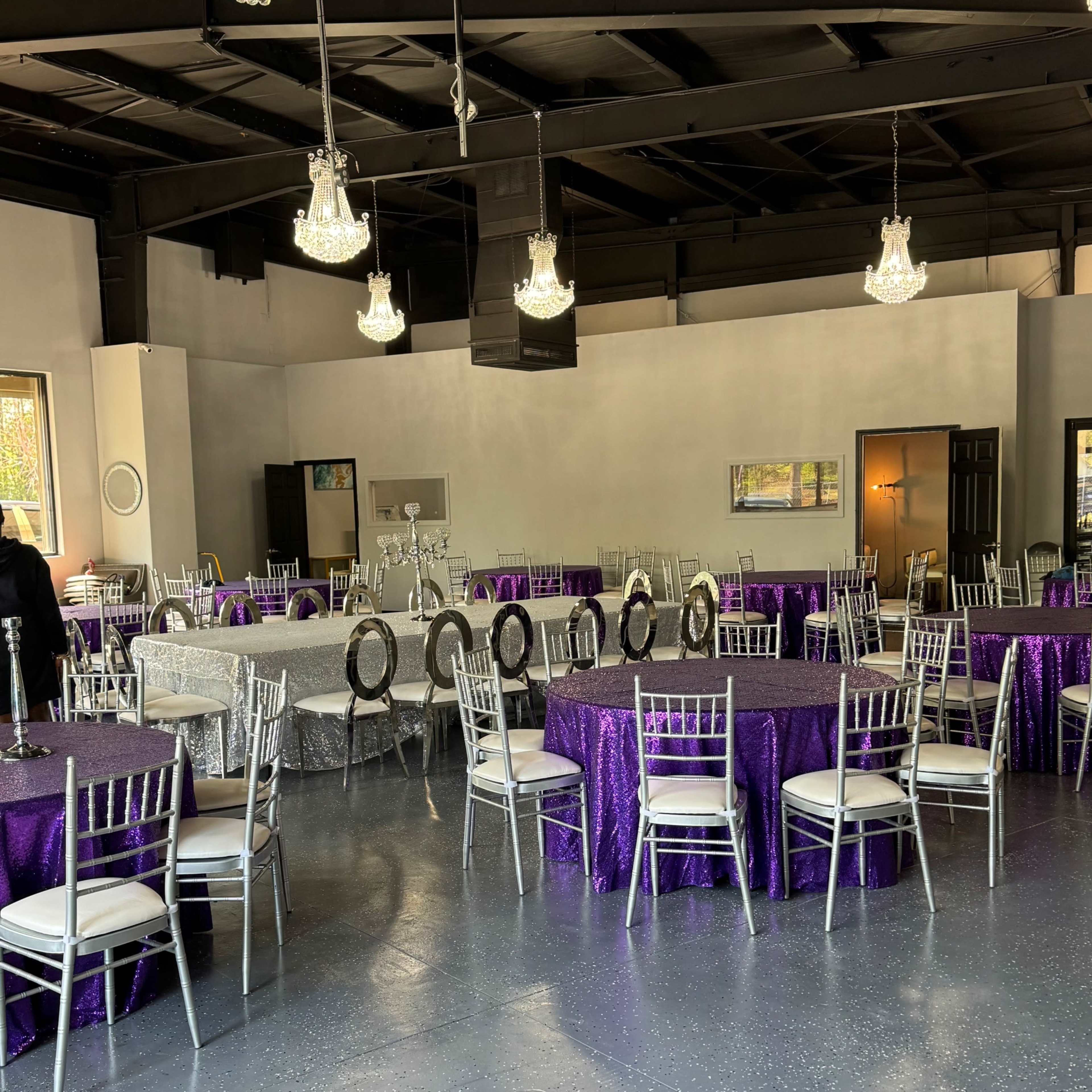 A brightly lit event space with silver chiavari chairs and tables covered in purple tablecloths.