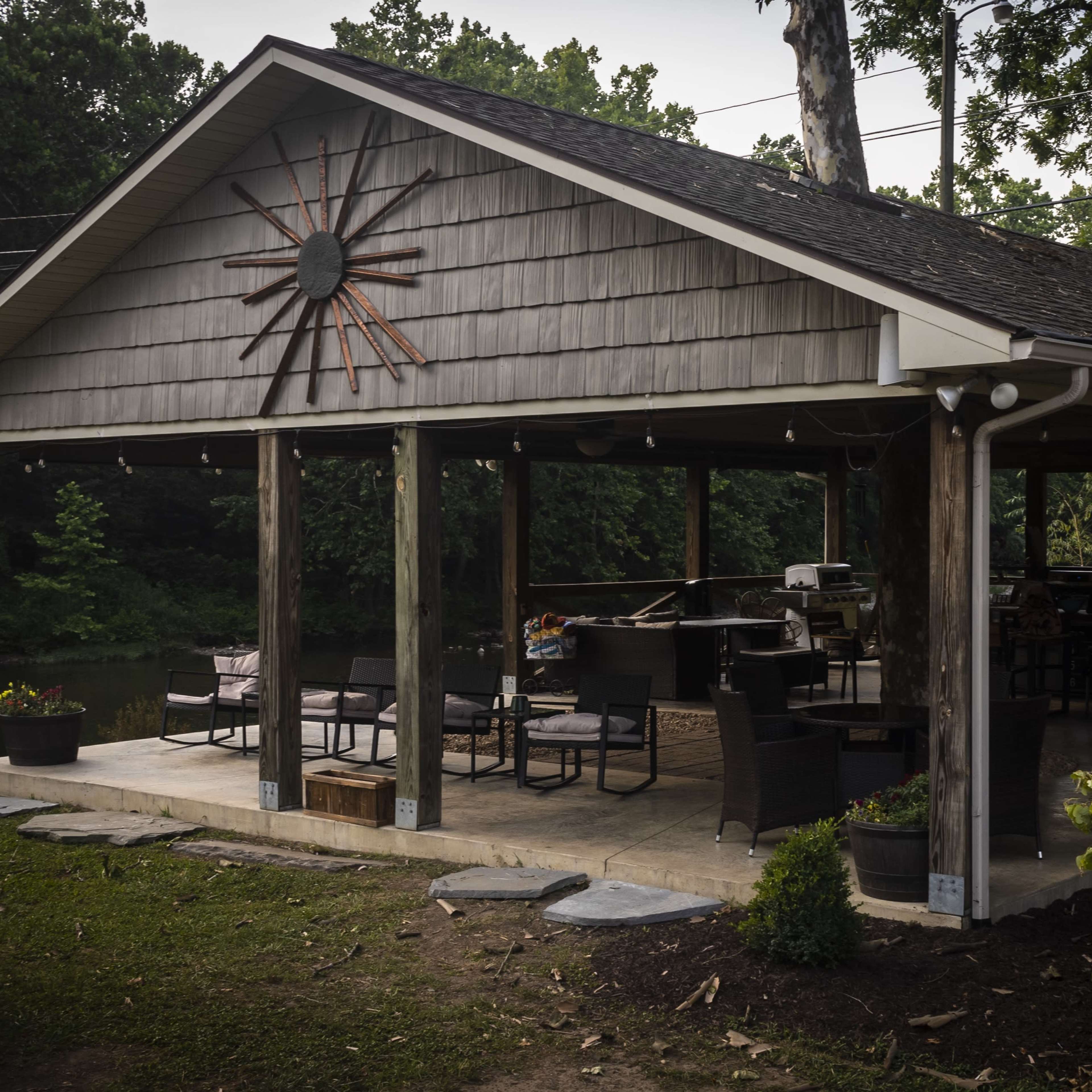 A wooden pavilion with a stone patio, featuring seating, a grill, and a large sunburst decoration on the wall, situated near a body of water surrounded by trees.
