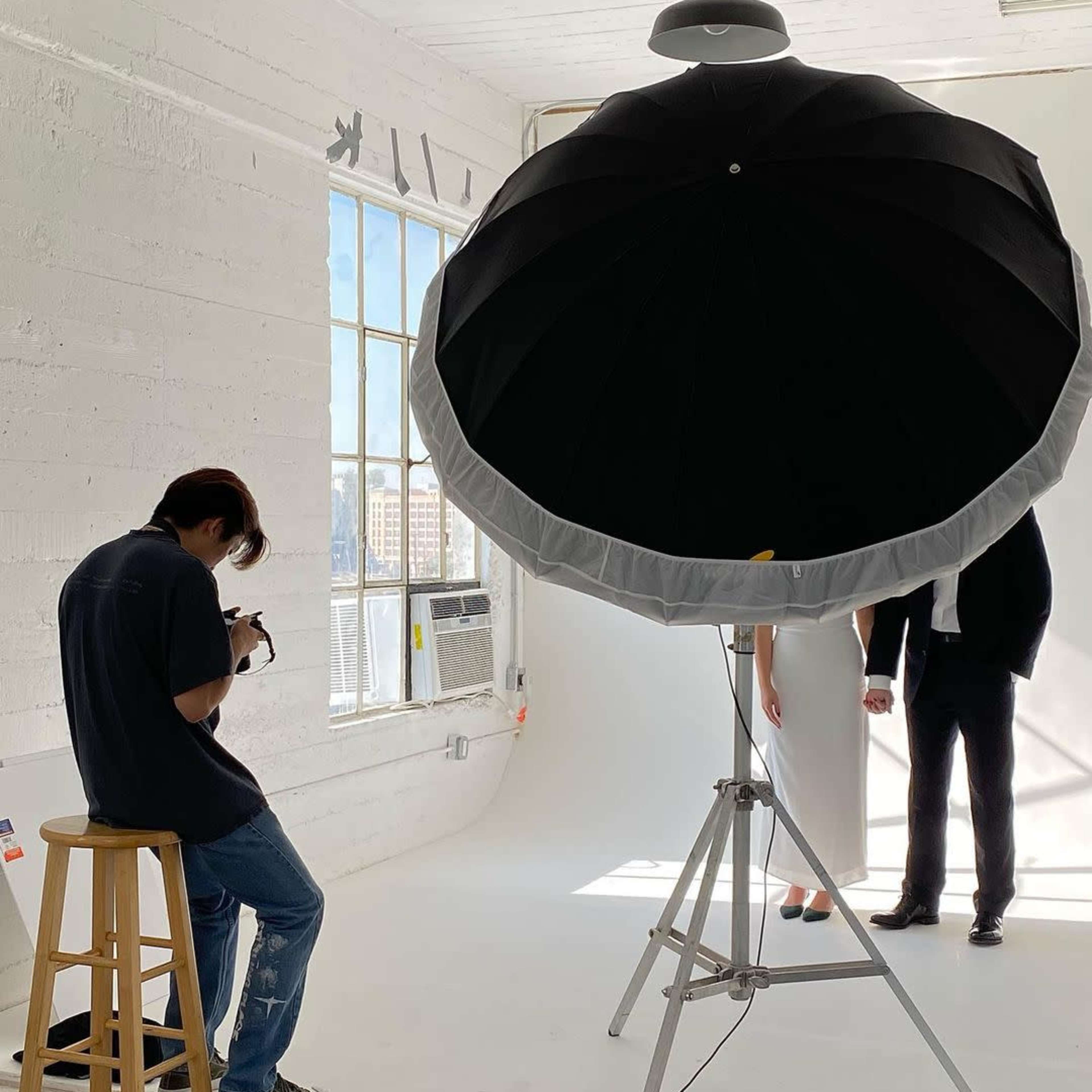 A photographer sits on a stool in a bright studio, capturing images of two models standing against a white backdrop, with a large light umbrella set up nearby.