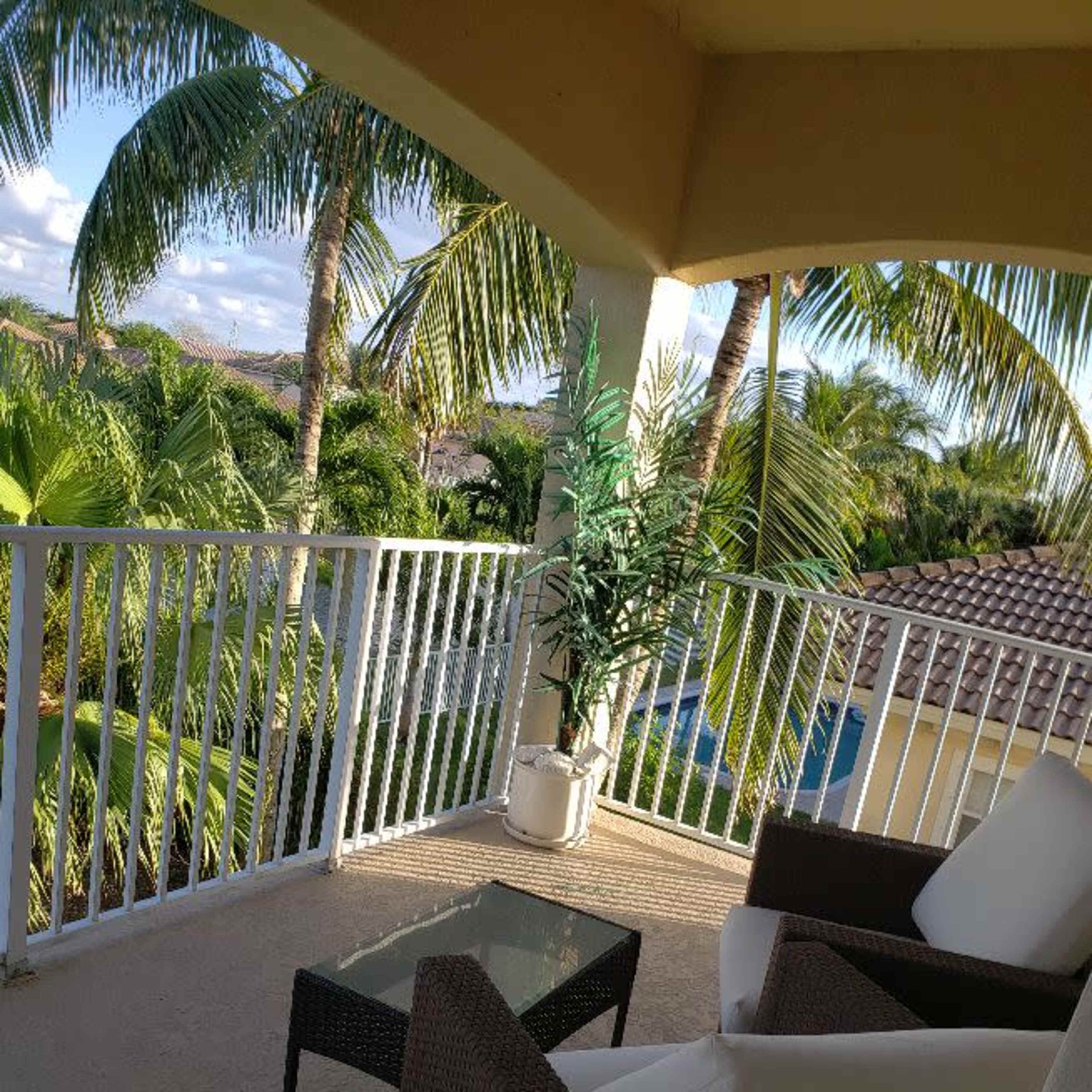 A balcony features a glass table and two wicker chairs surrounded by tall palm trees and lush greenery.