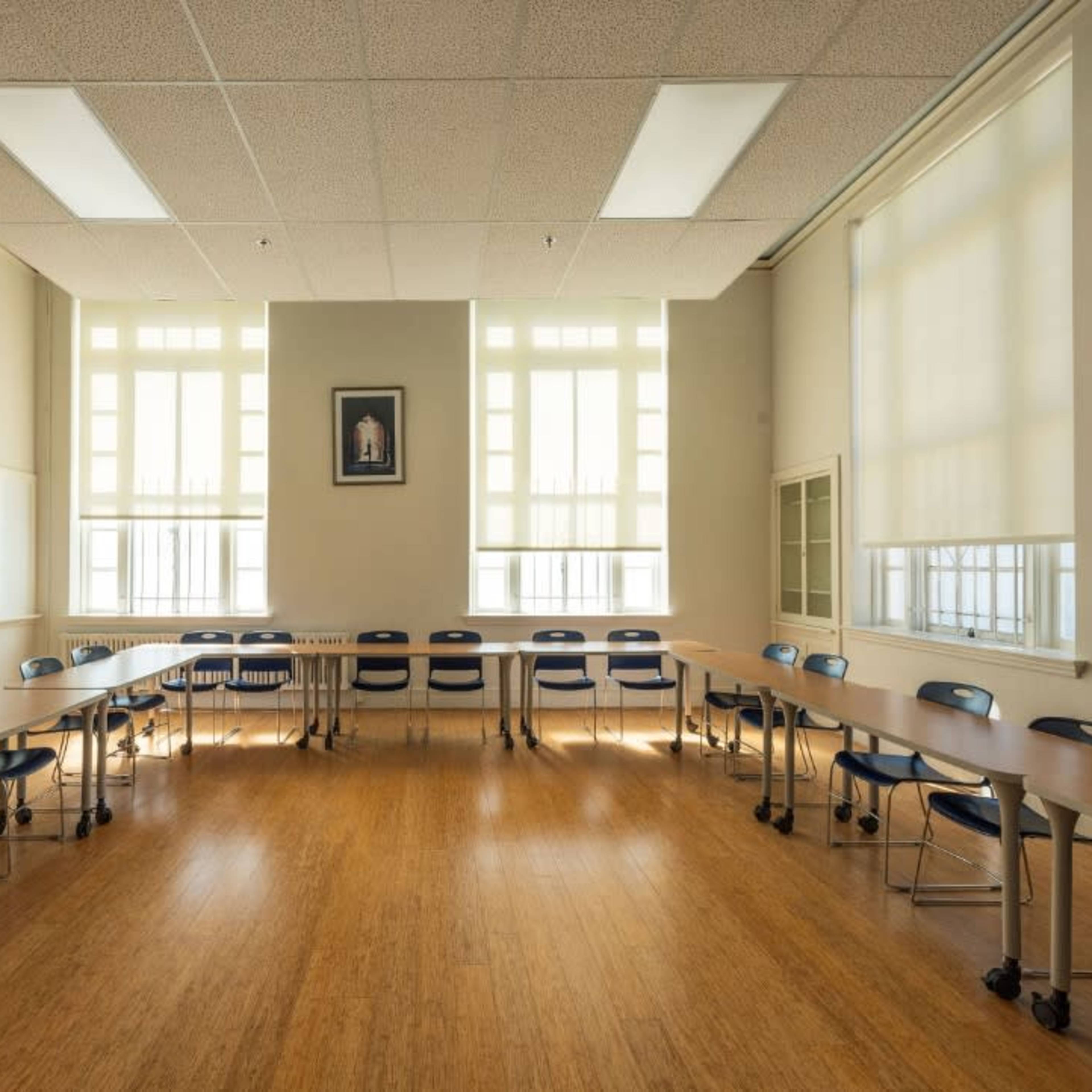 A spacious classroom is arranged with tables and chairs in a circular formation, illuminated by large windows.