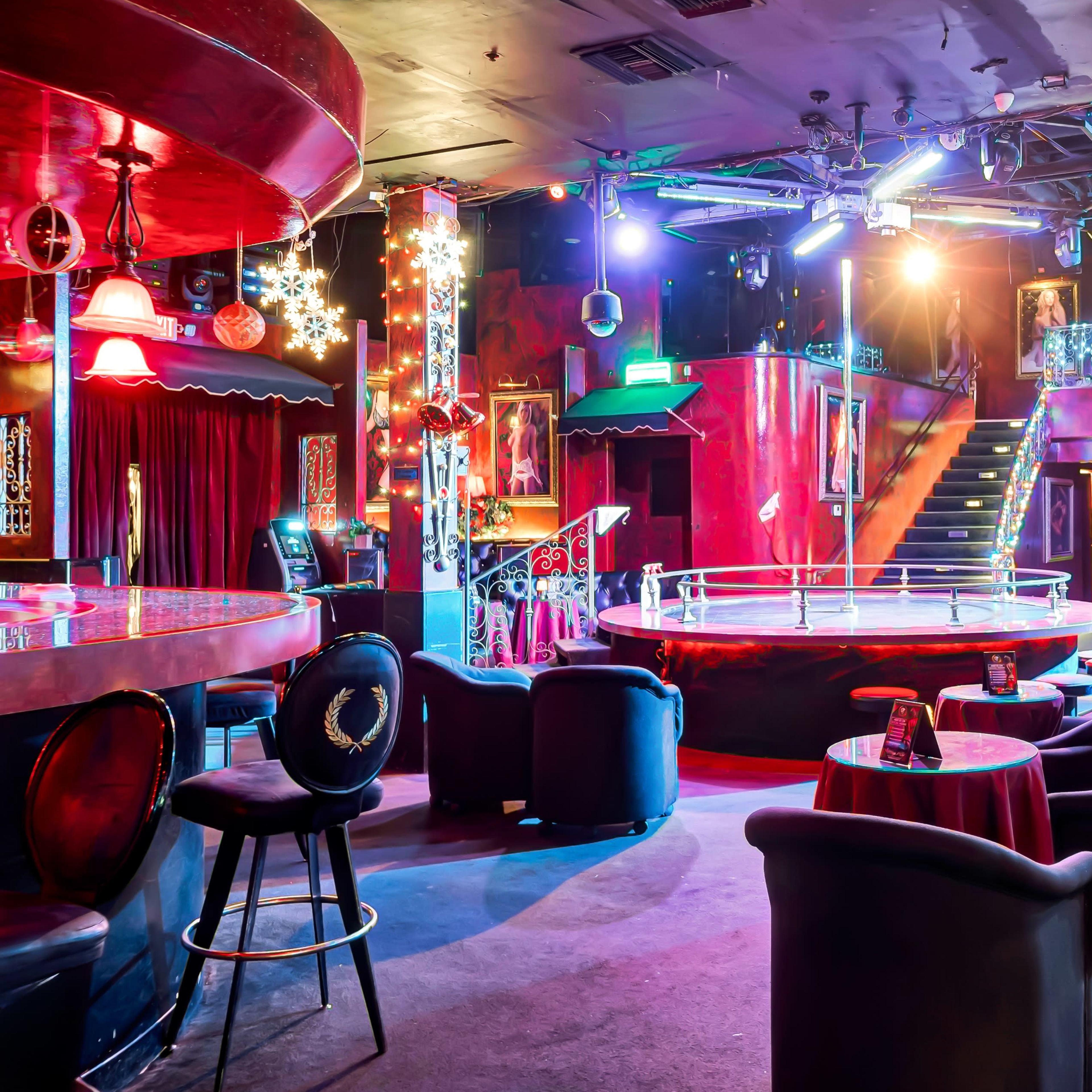 An empty nightclub interior featuring a circular performance stage, seating areas with dark chairs, and colorful lighting.