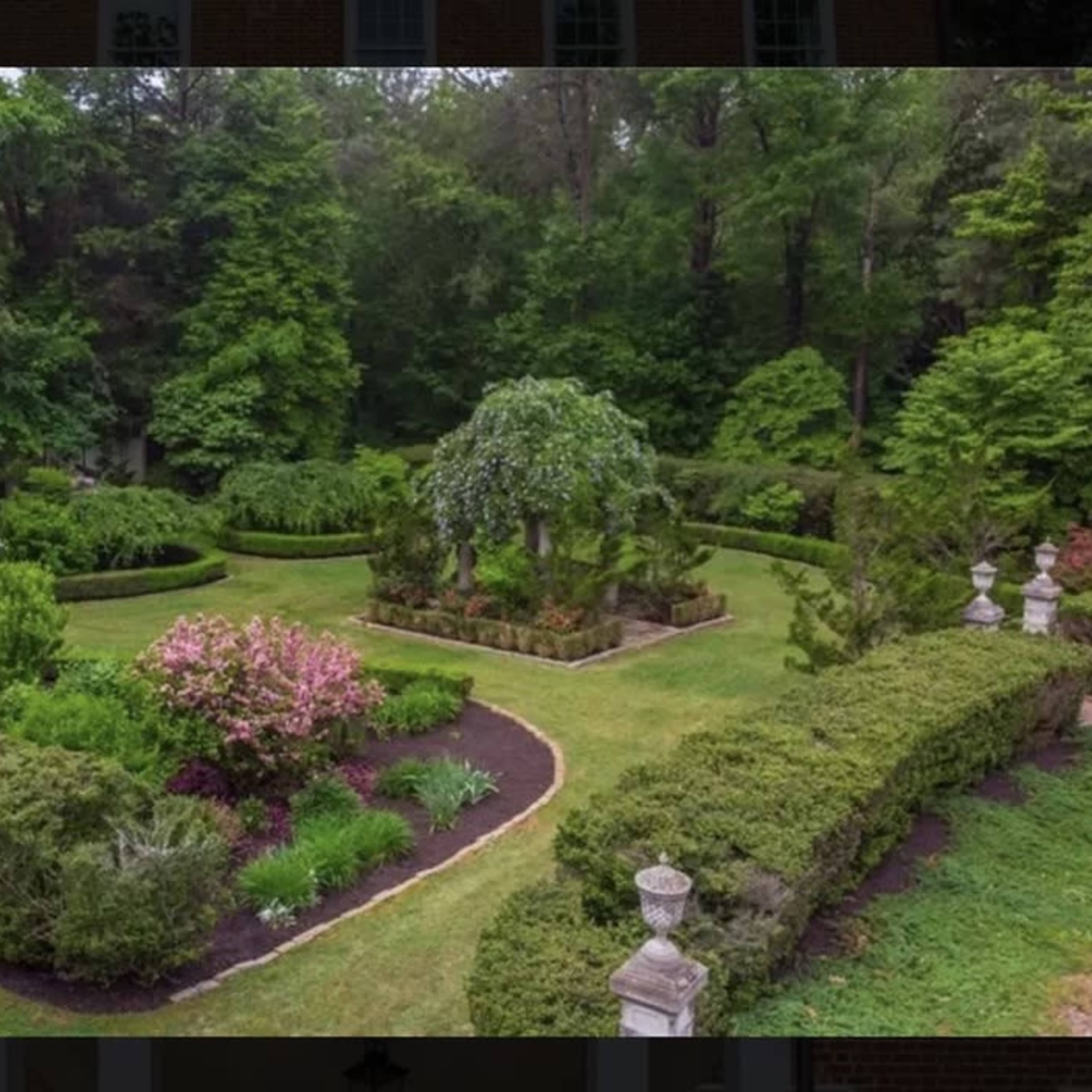 A well-maintained garden features neatly trimmed hedges, colorful flowerbeds, and a central tree surrounded by manicured paths.