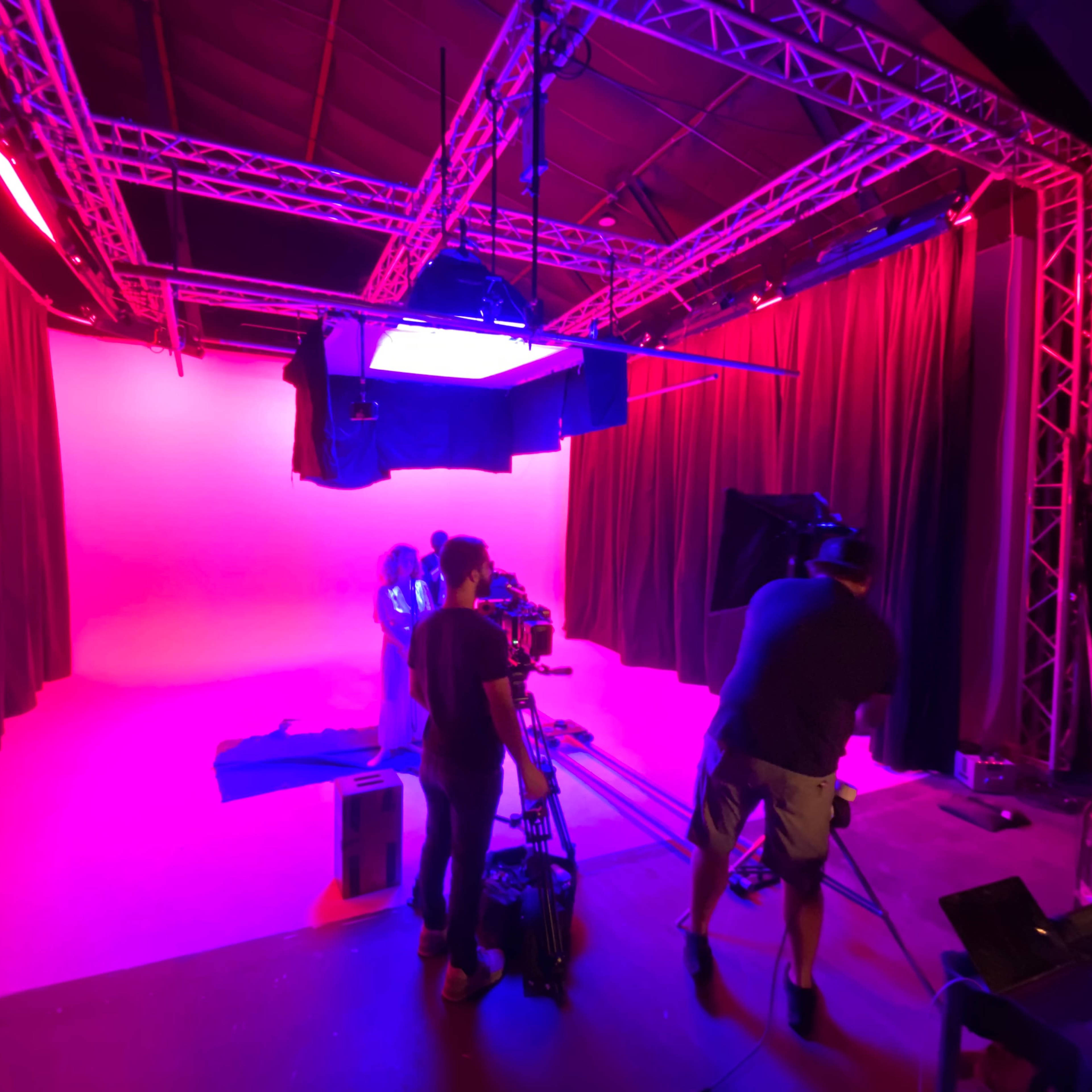 A film crew operates equipment in a brightly lit studio with pink and purple background lighting.