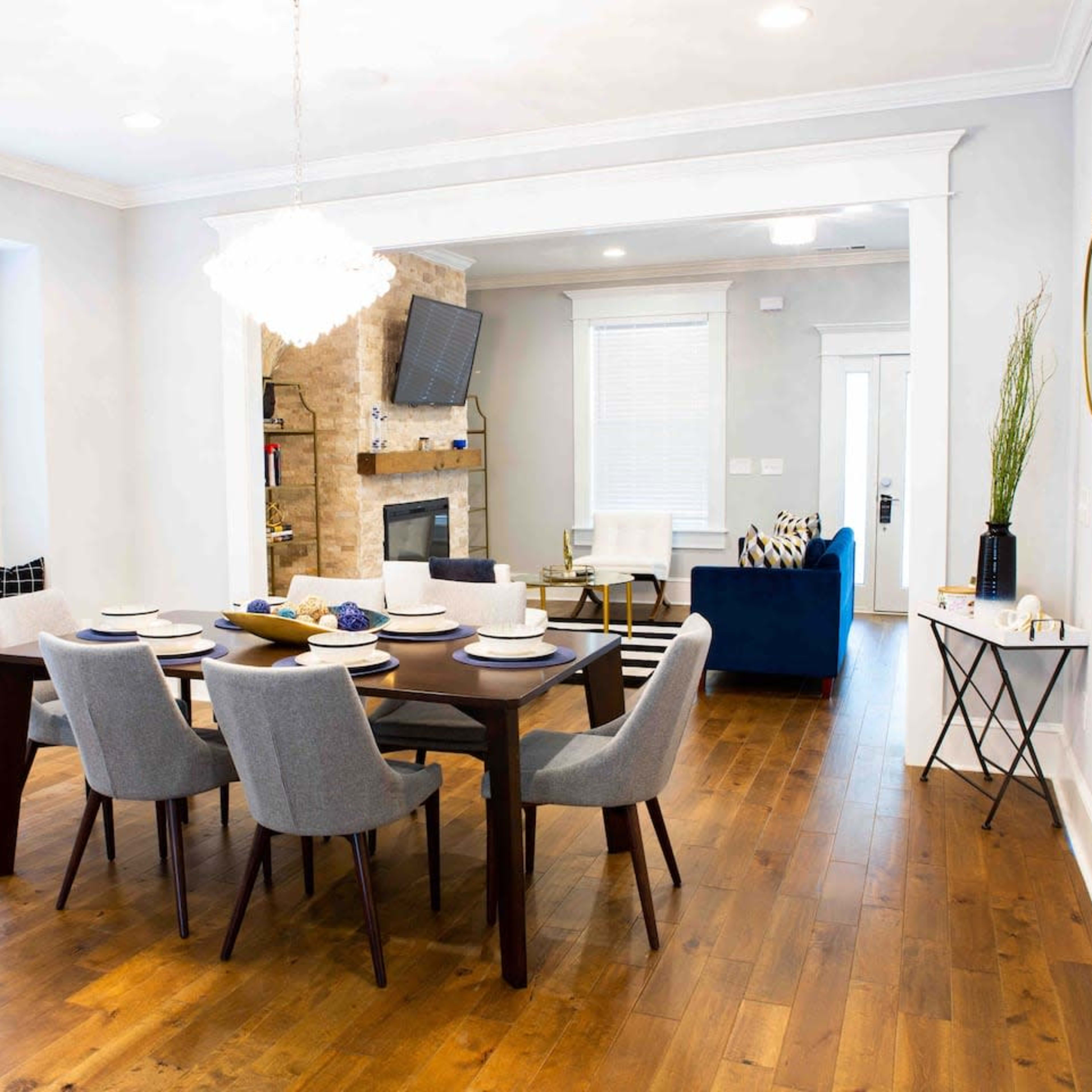 A dining area features a rectangular table set for eight, surrounded by gray chairs, with a stylish light fixture overhead and a cozy living room partially visible in the background.