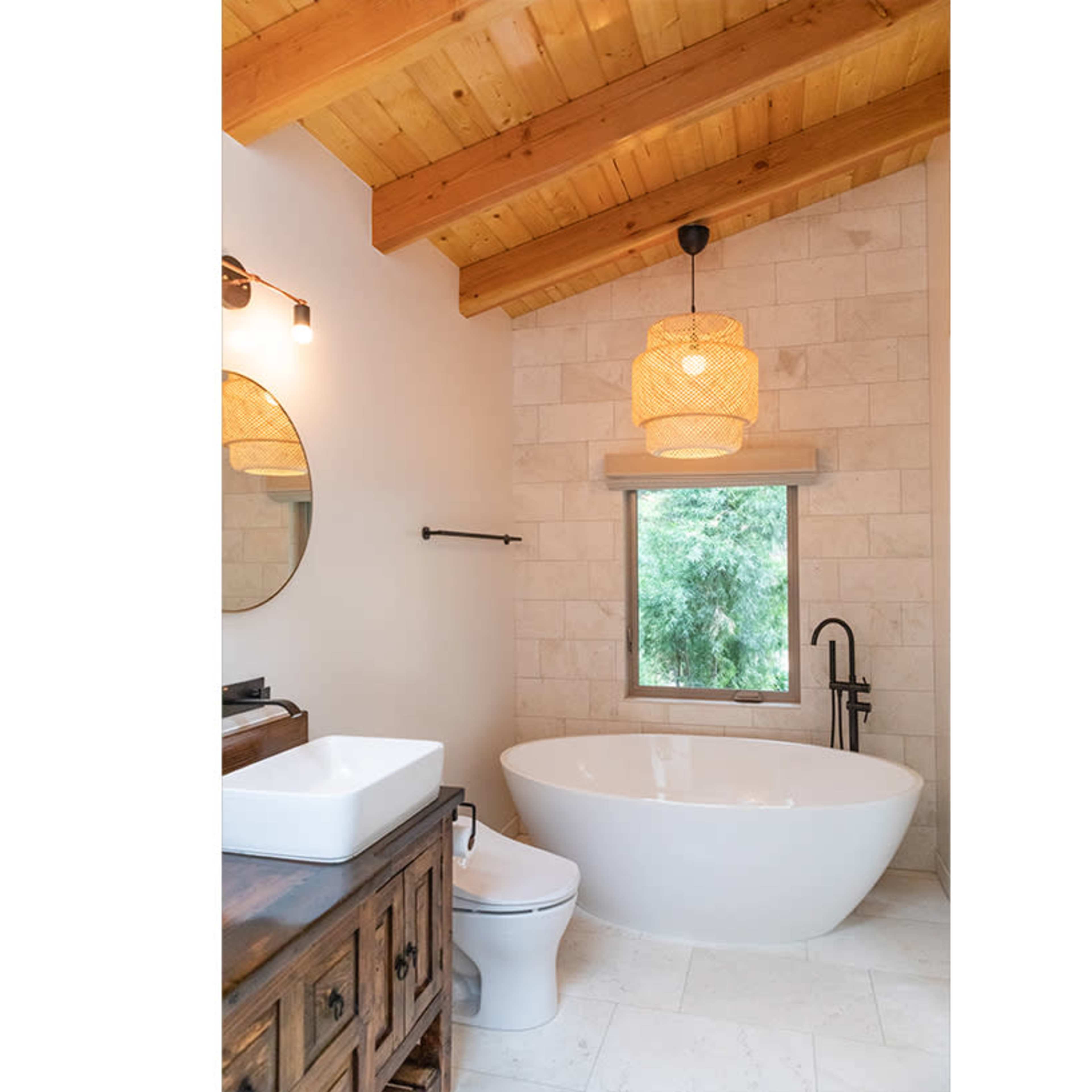 A modern bathroom with a freestanding bathtub, a wooden vanity, a round mirror, and a window overlooking greenery, all complemented by wooden ceiling beams and stone tiles.