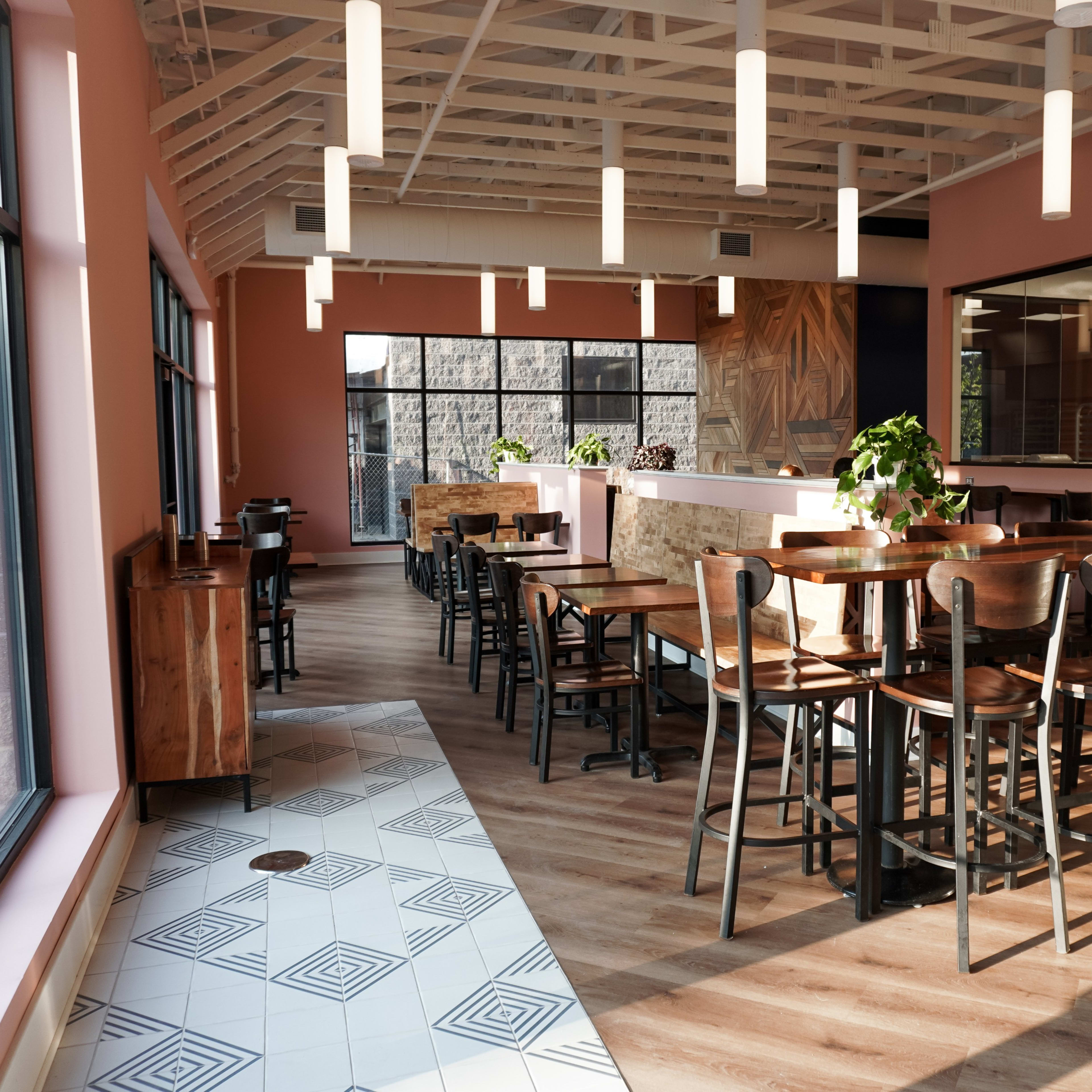A modern restaurant interior with wooden tables and chairs, large windows, and a light pink and wood color scheme.