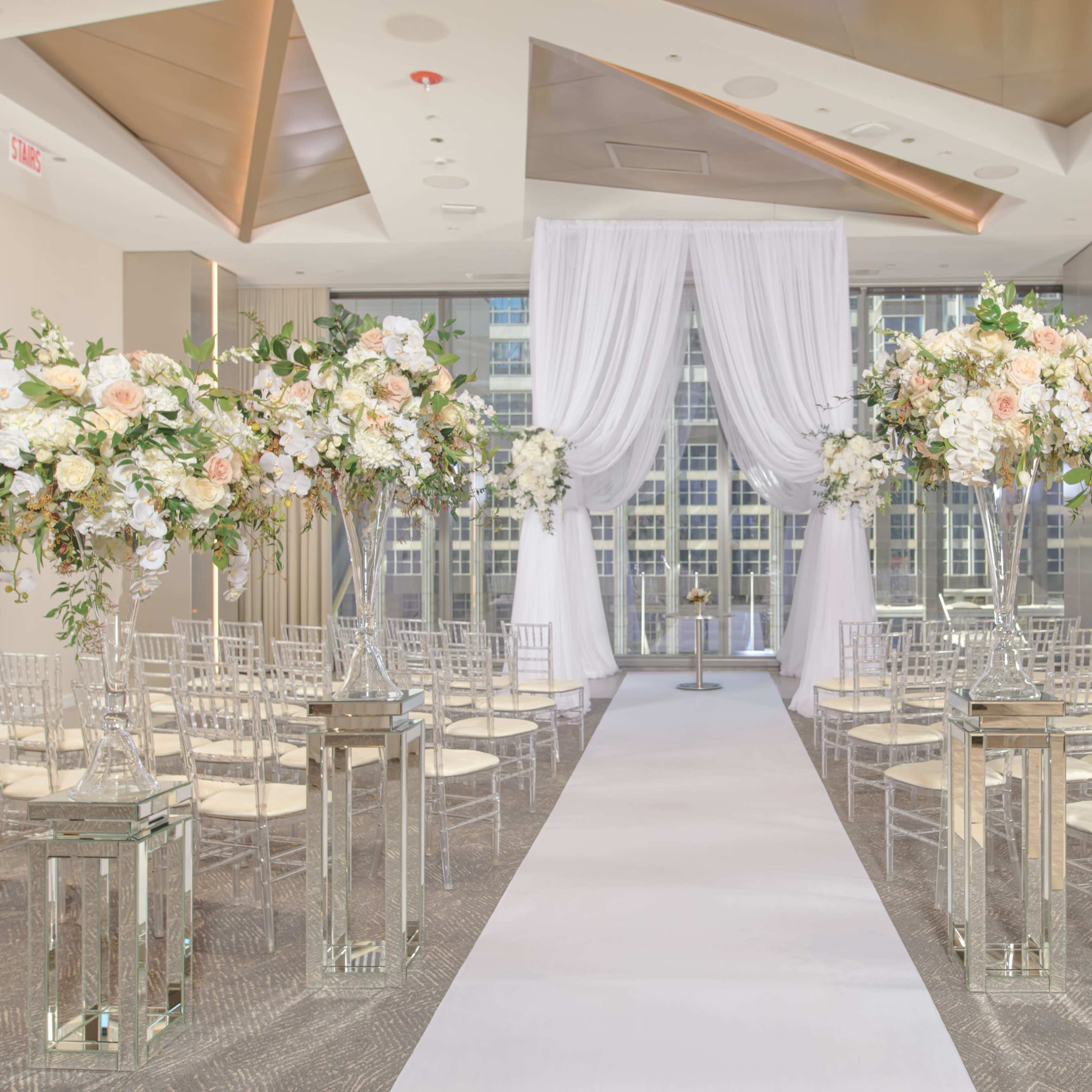 A wedding ceremony setup features rows of chairs aligned on a white aisle, flanked by tall floral arrangements and decorative lanterns, with sheer drapes framing a backdrop.
