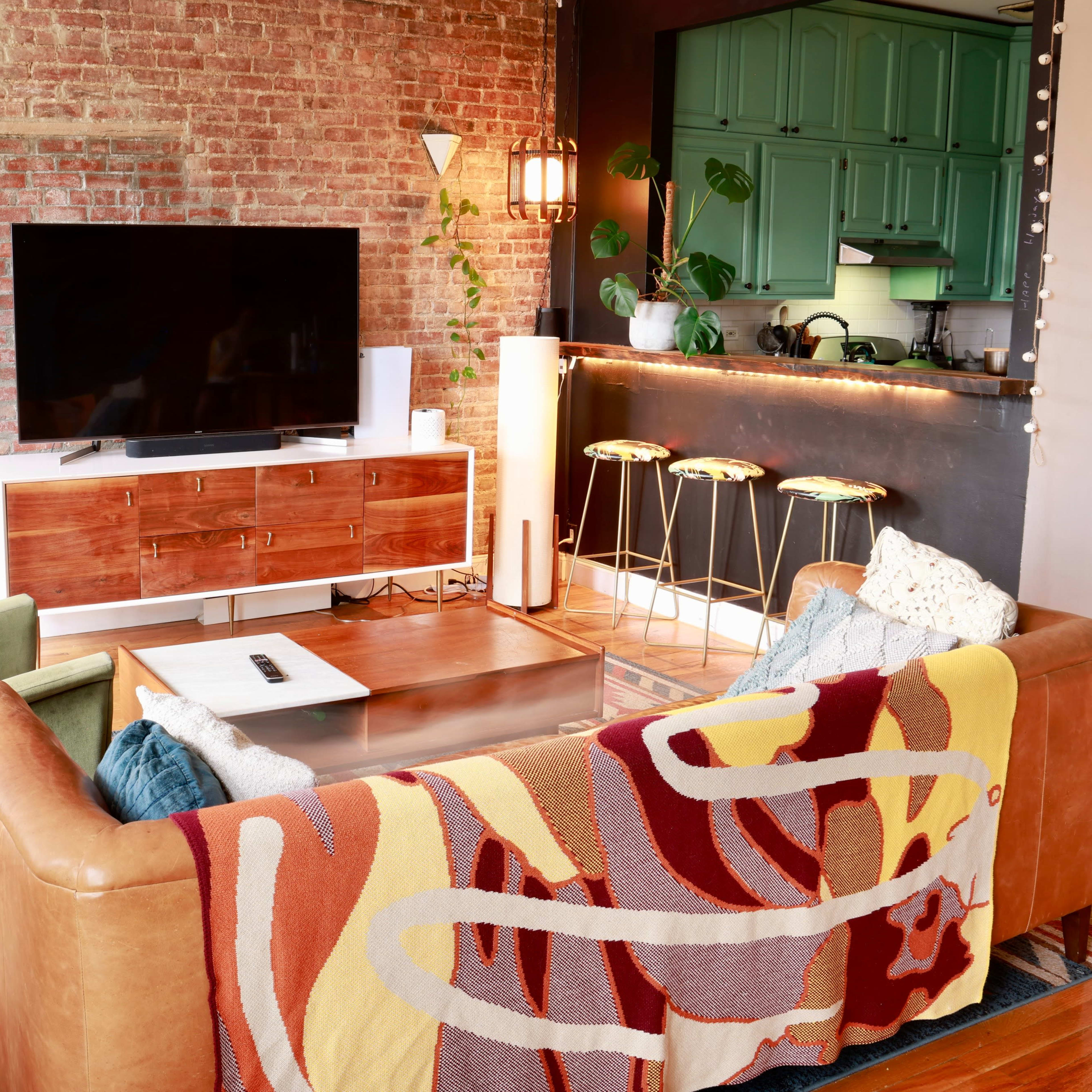 A cozy living room with exposed brick walls, a large television, and a kitchen area featuring green cabinets and bar stools.