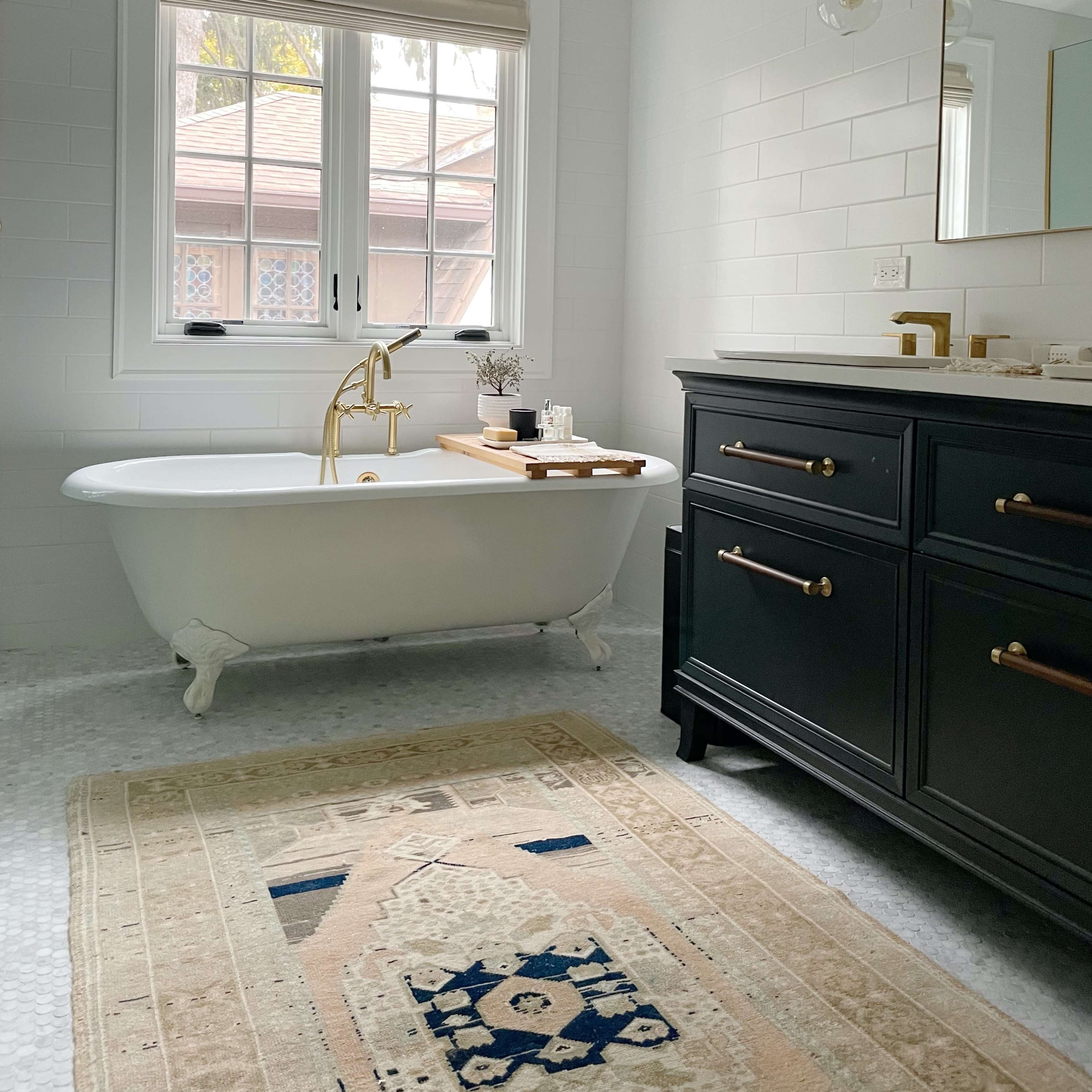 A bright bathroom featuring a freestanding bathtub, a black vanity with gold hardware, and a patterned area rug on a stone-tiled floor.