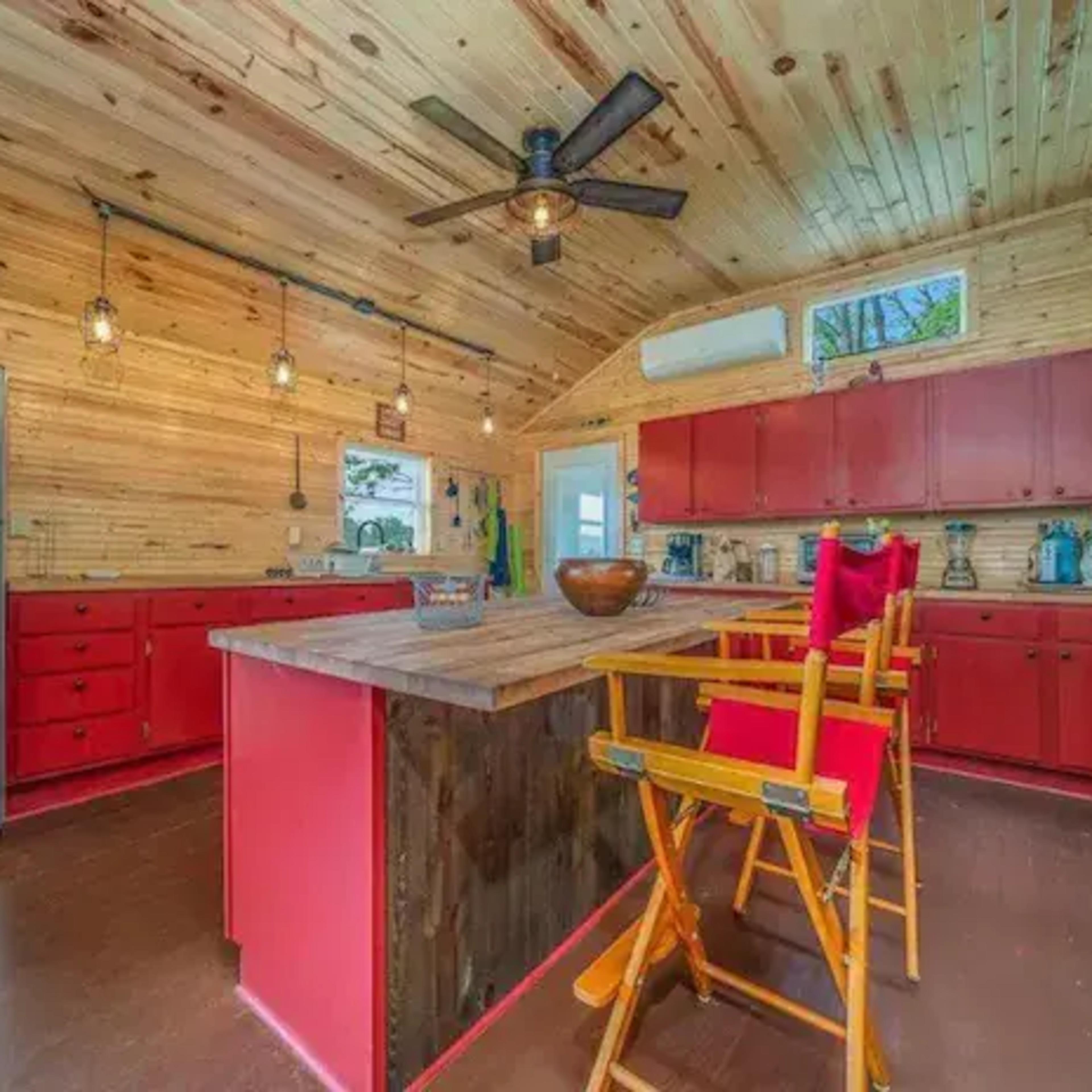 A rustic kitchen with wooden walls and ceiling, featuring red cabinets, stainless steel appliances, and a wooden island with tall chairs.
