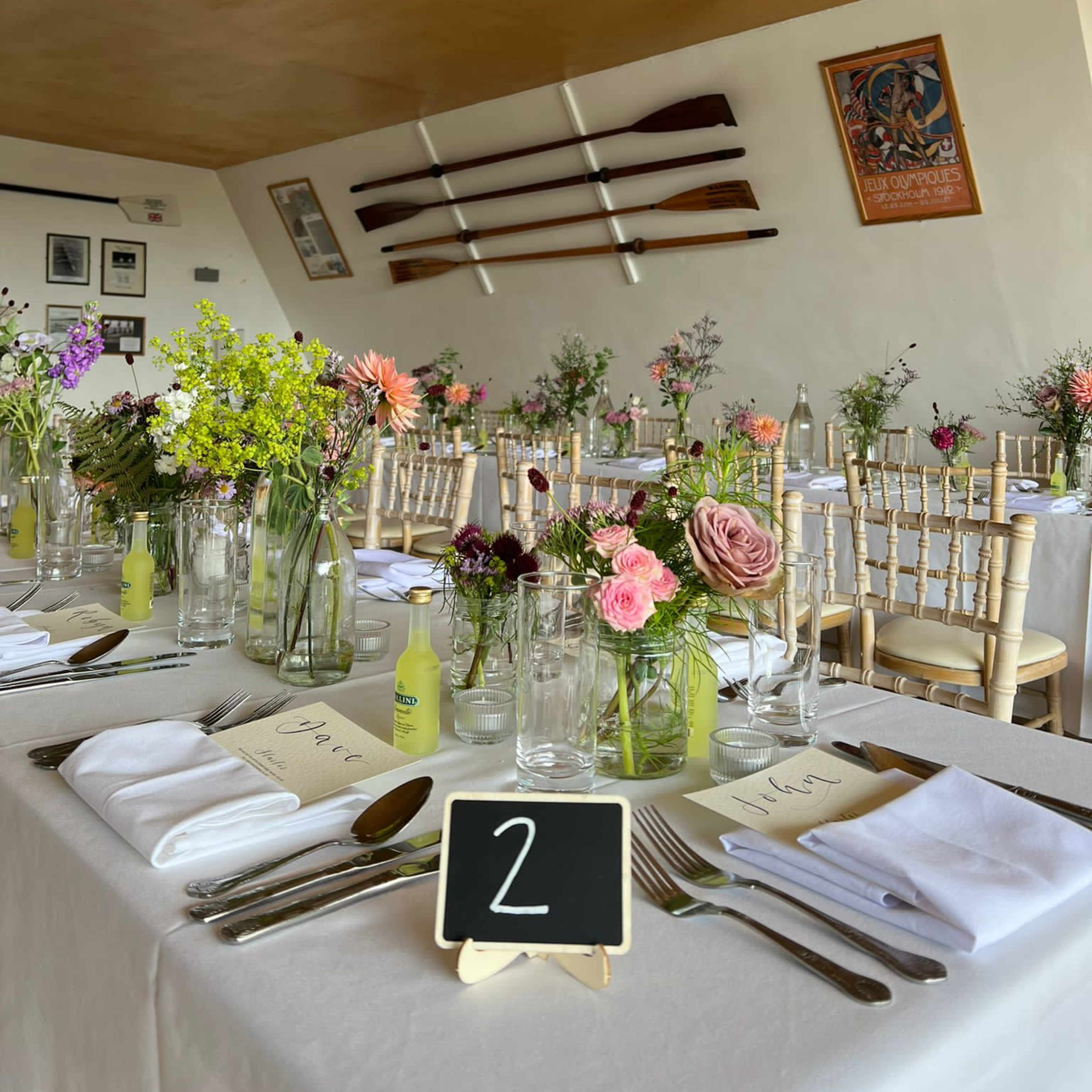 A dining area is set with tables neatly arranged with floral centerpieces and numbered place cards, while oars are displayed on the wall.