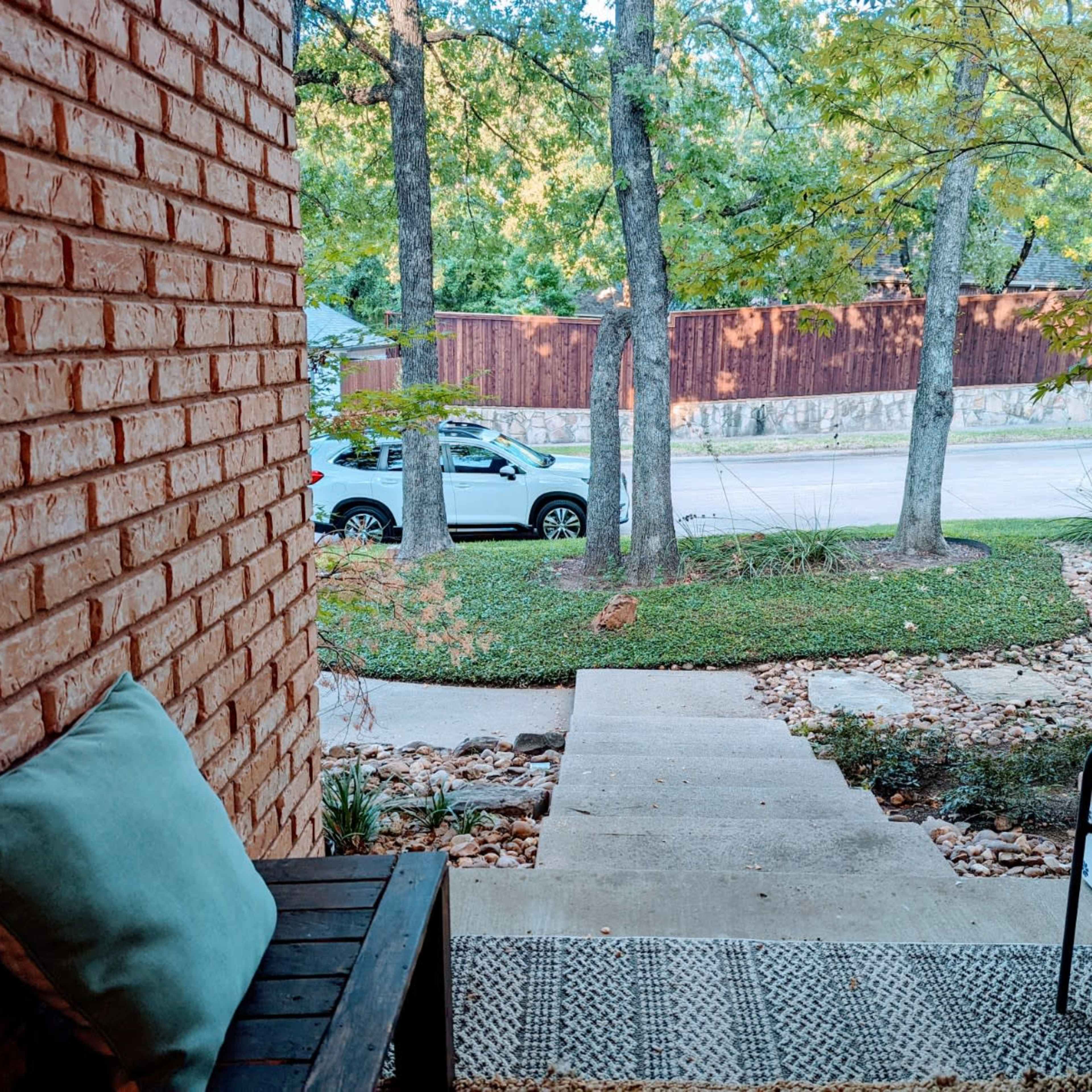 A view from a porch with brick walls, a bench, and a pathway leading to a street lined with trees and a parked white vehicle.