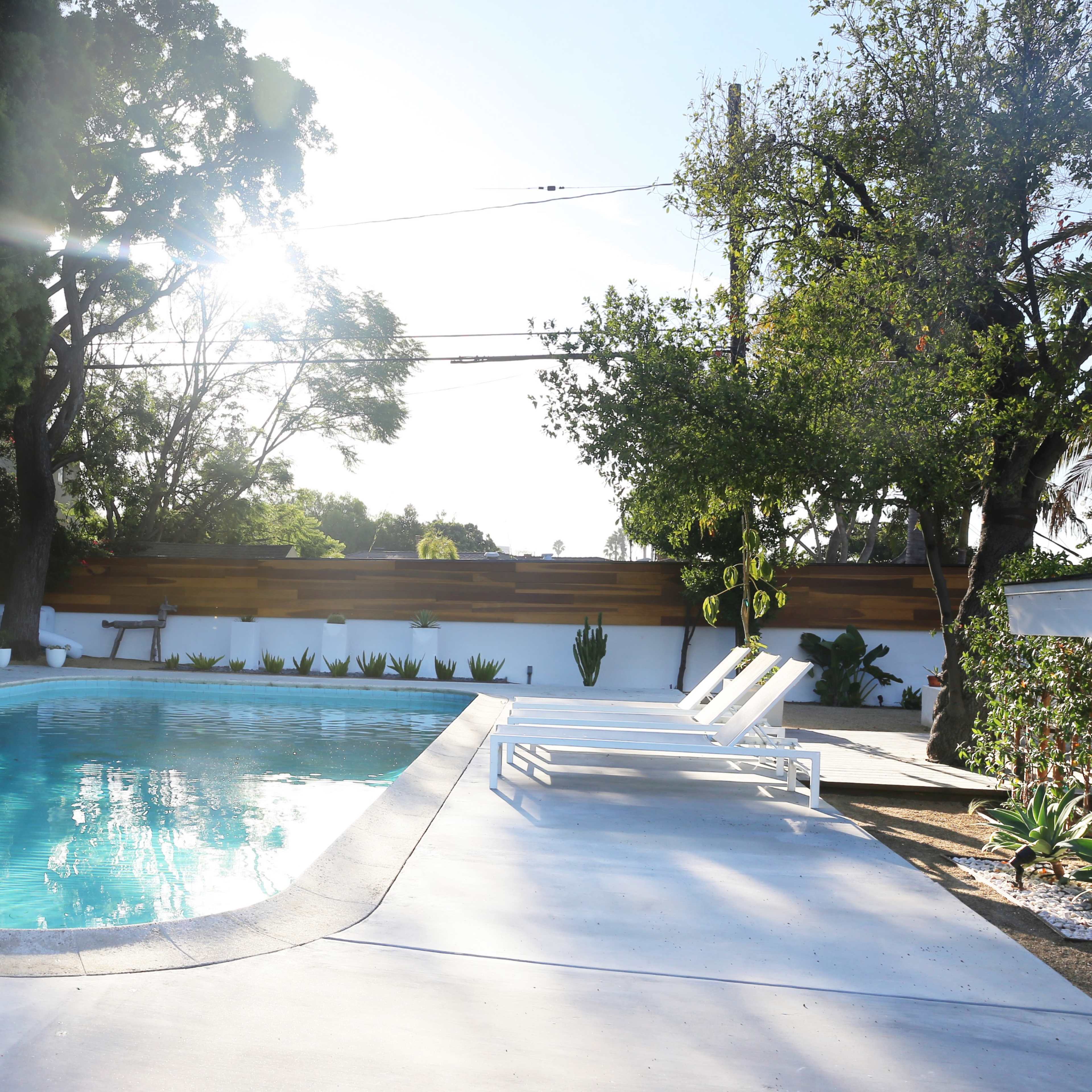 A clear swimming pool is surrounded by sun loungers and landscaped greenery in a bright outdoor setting.