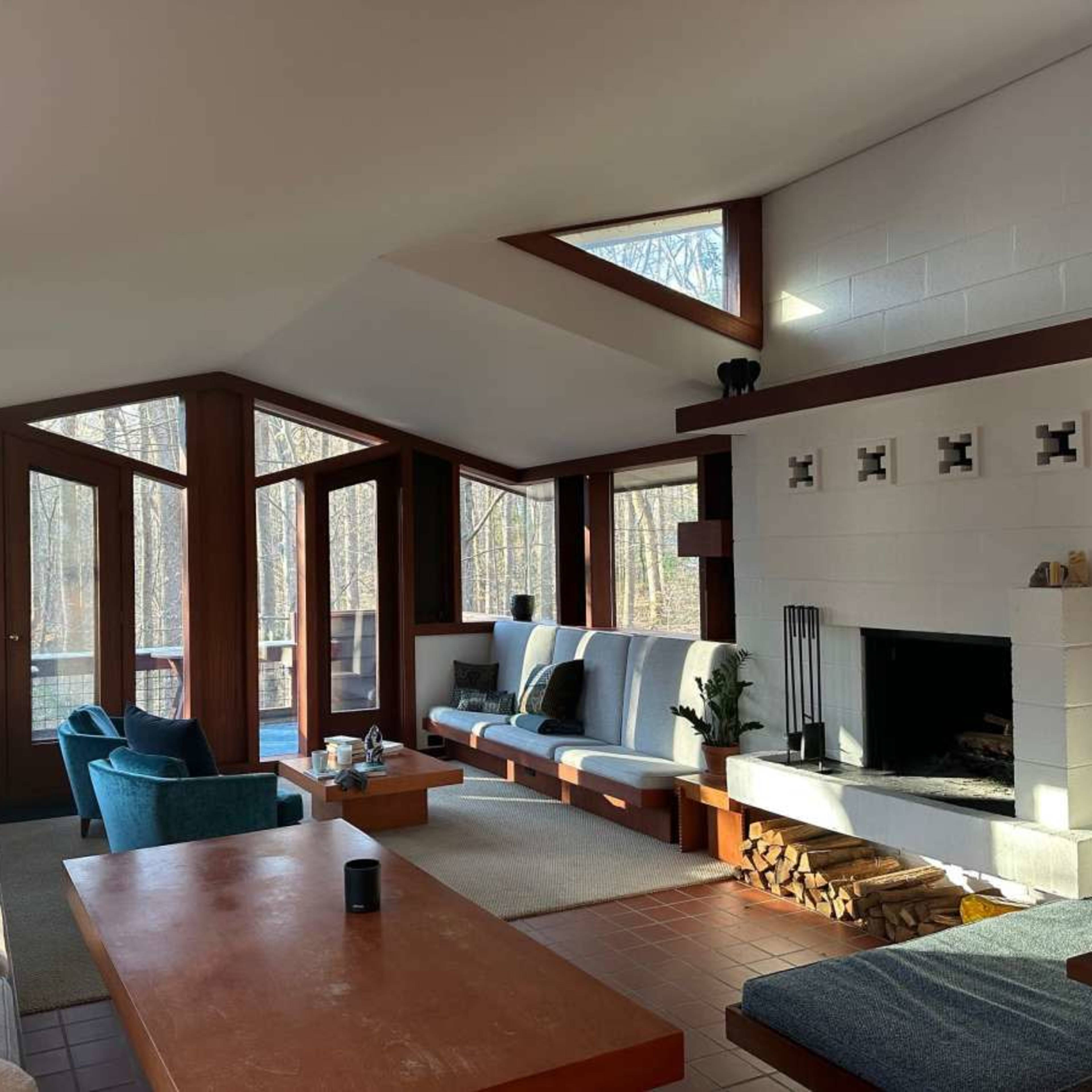 A modern living room with large windows overlooking a wooded area, featuring a fireplace, comfortable seating, and wooden furniture.