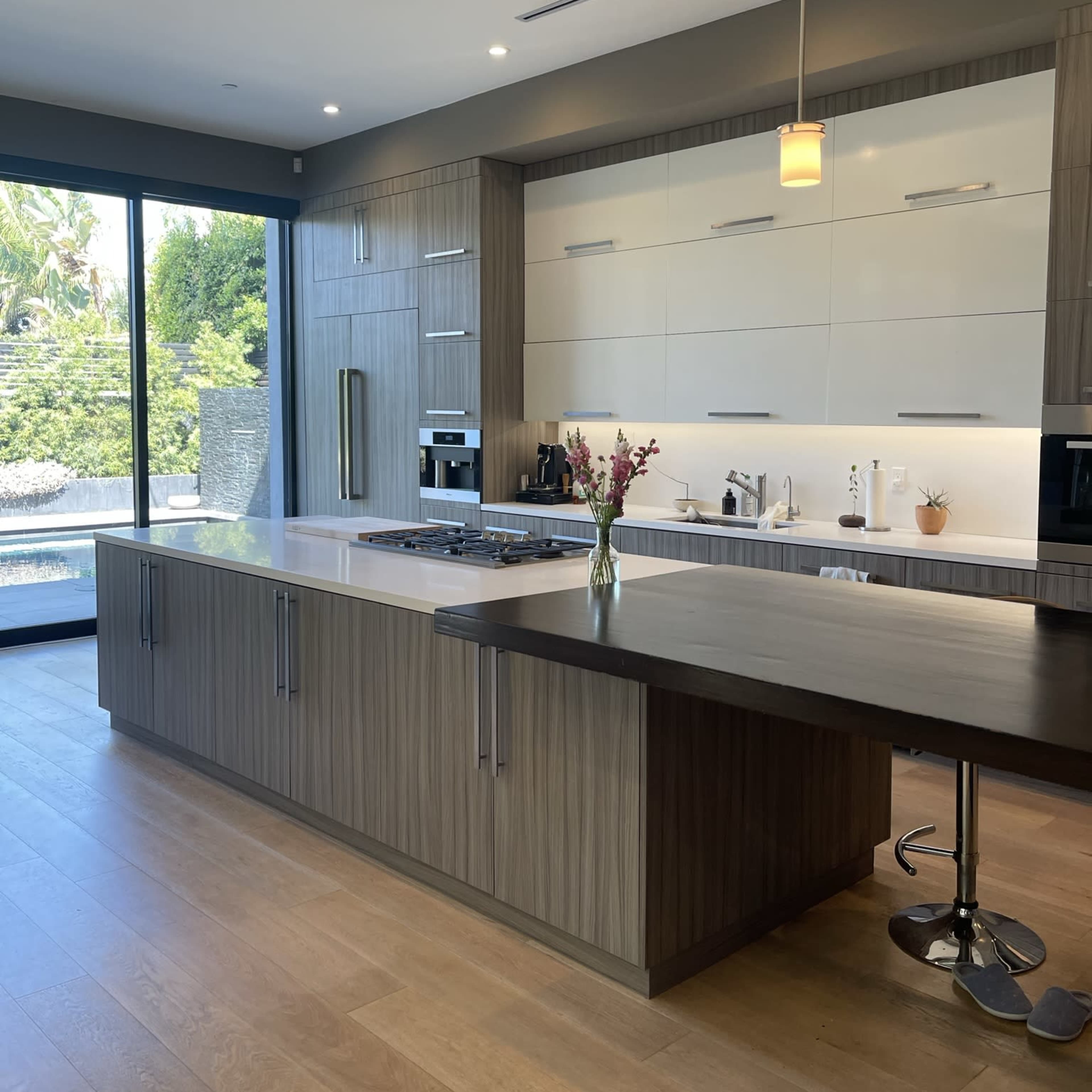A modern kitchen features a central island with a stovetop, sleek cabinetry, and large windows overlooking a garden.