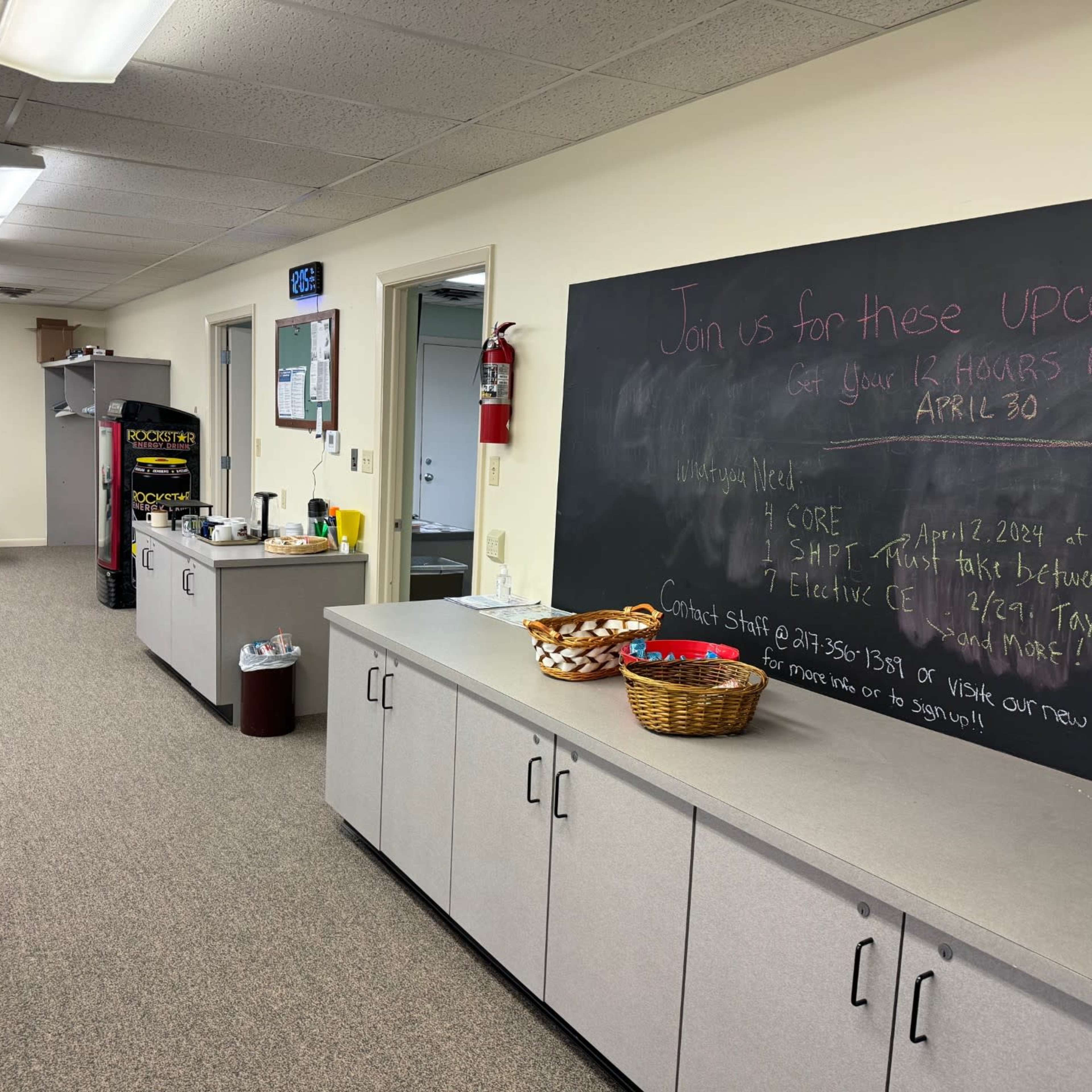 A bright room features a snack area, a chalkboard with upcoming events, and empty tables along one wall.