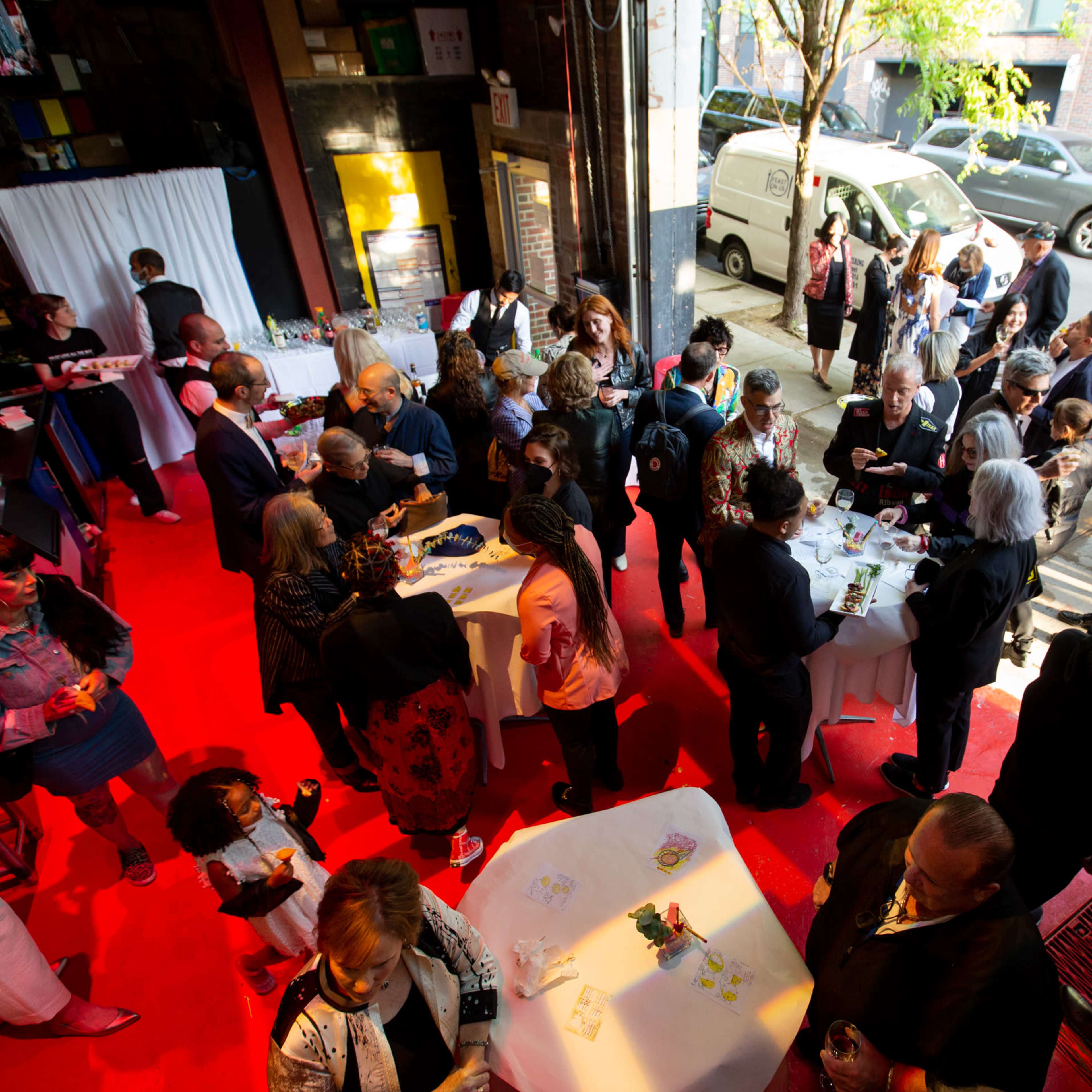 A large group of people mingles at an event in an urban space with red lighting and tables set up for refreshments.