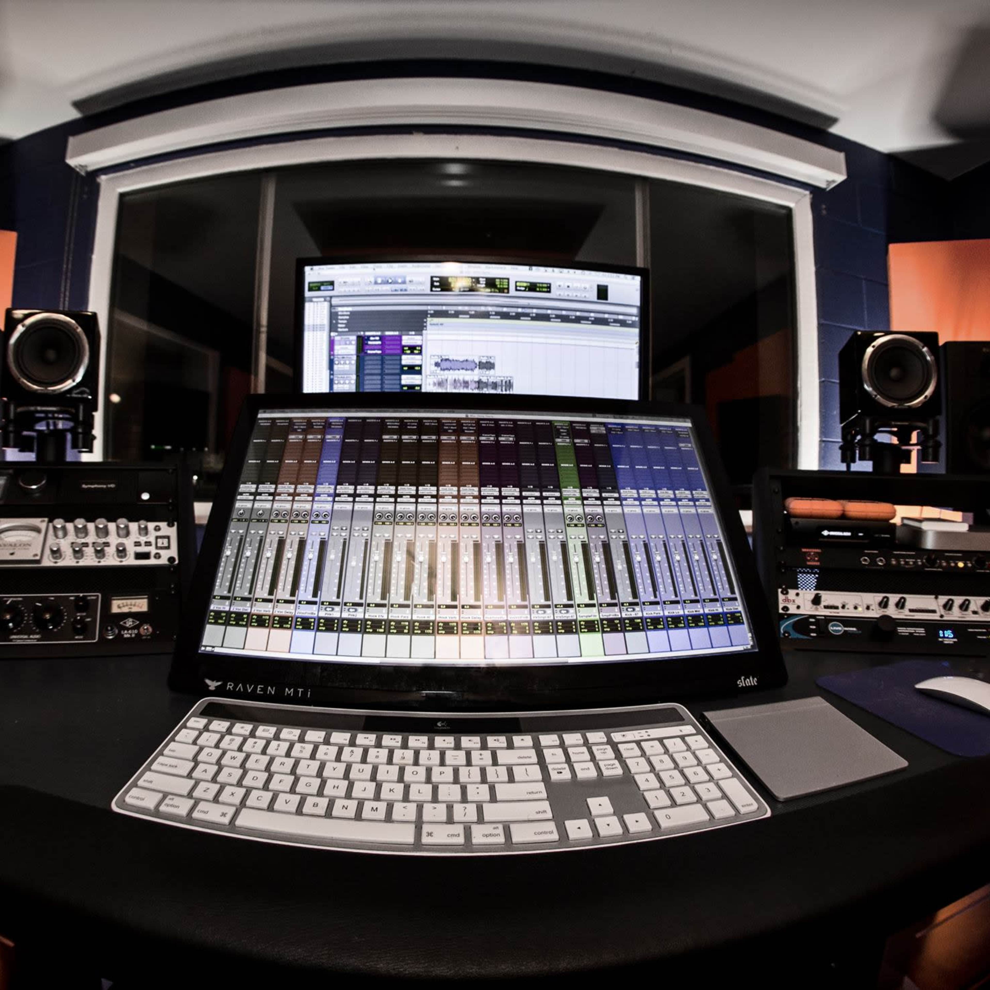 A modern audio recording studio with a large mixing console, computer monitors, and studio speakers arranged around a central workstation.