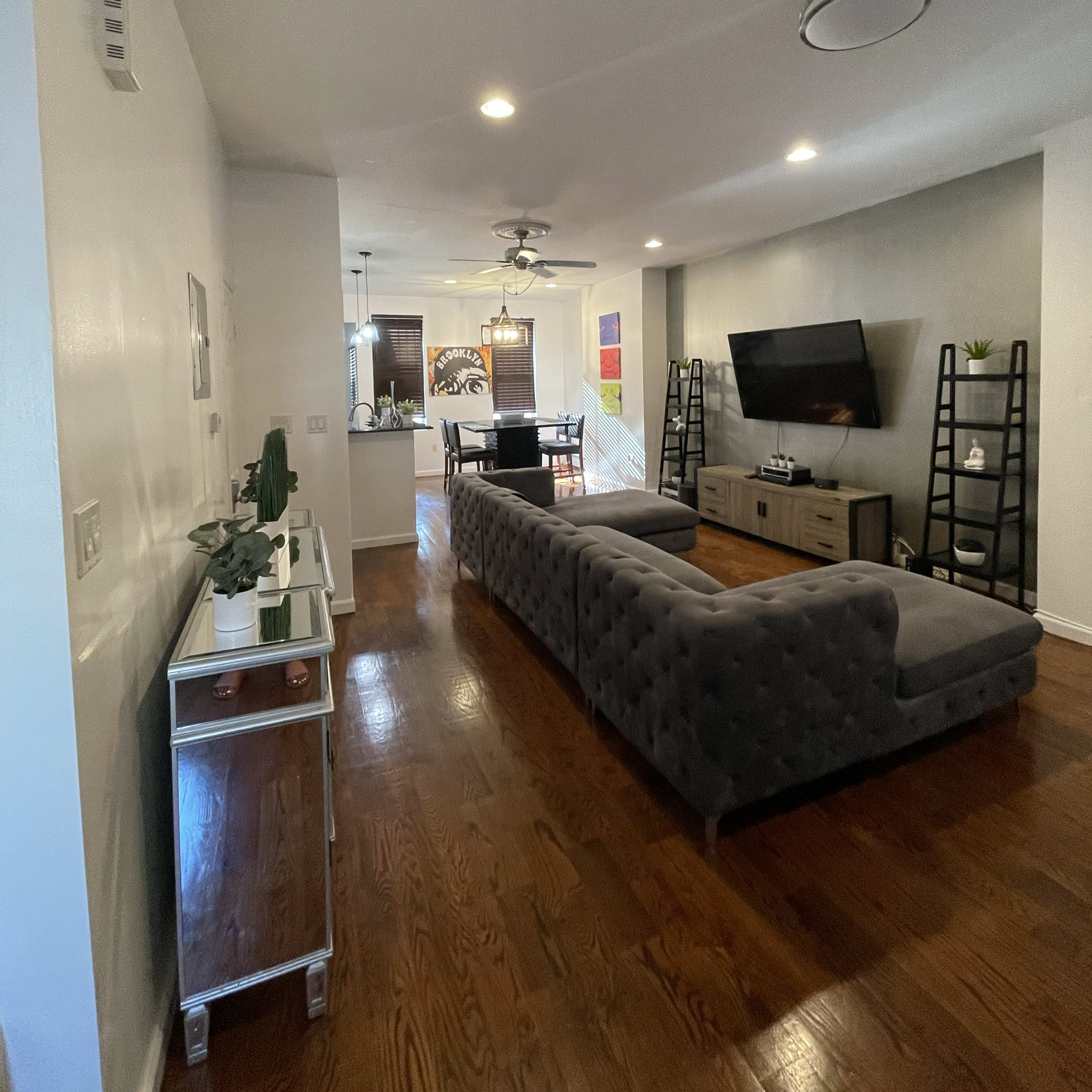 A modern living room features a gray sectional sofa, wooden flooring, and a television mounted on the wall, with a dining area visible in the background.