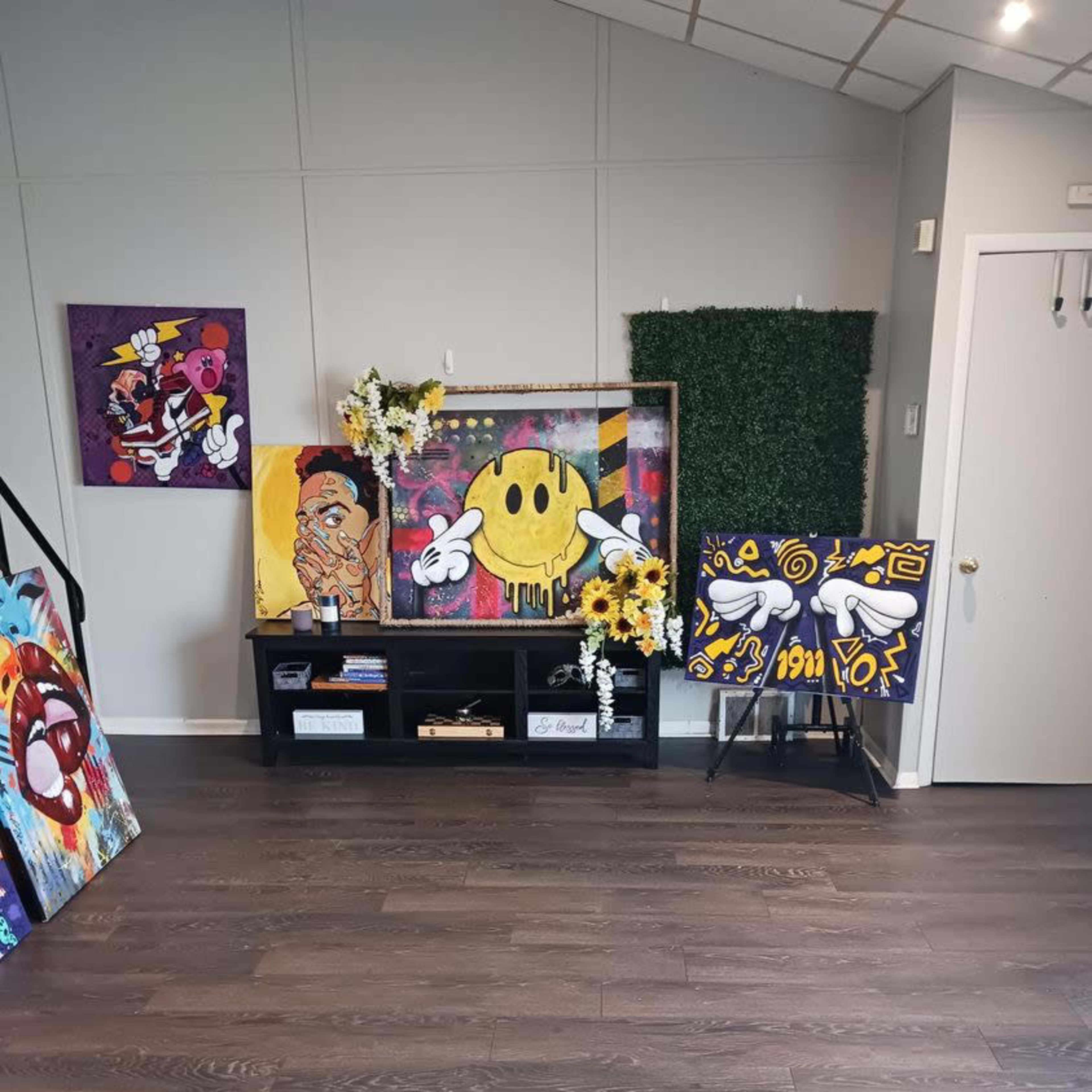 An art gallery interior featuring several colorful paintings, a shelf with decorative flowers, and a green wall panel.