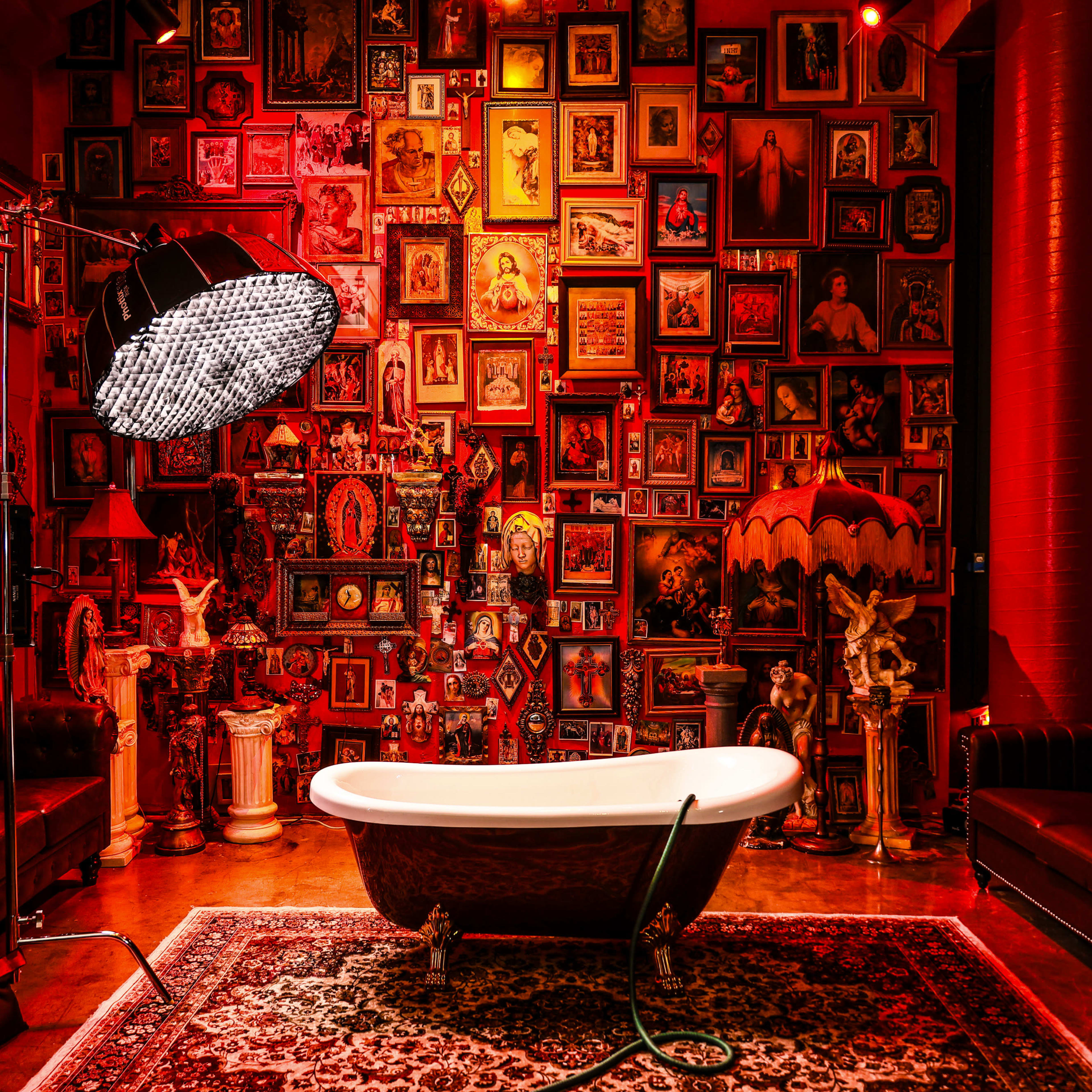 A luxurious bathroom featuring a vintage clawfoot tub, surrounded by an ornate wall filled with framed portraits and decorative objects, illuminated by a large studio light.