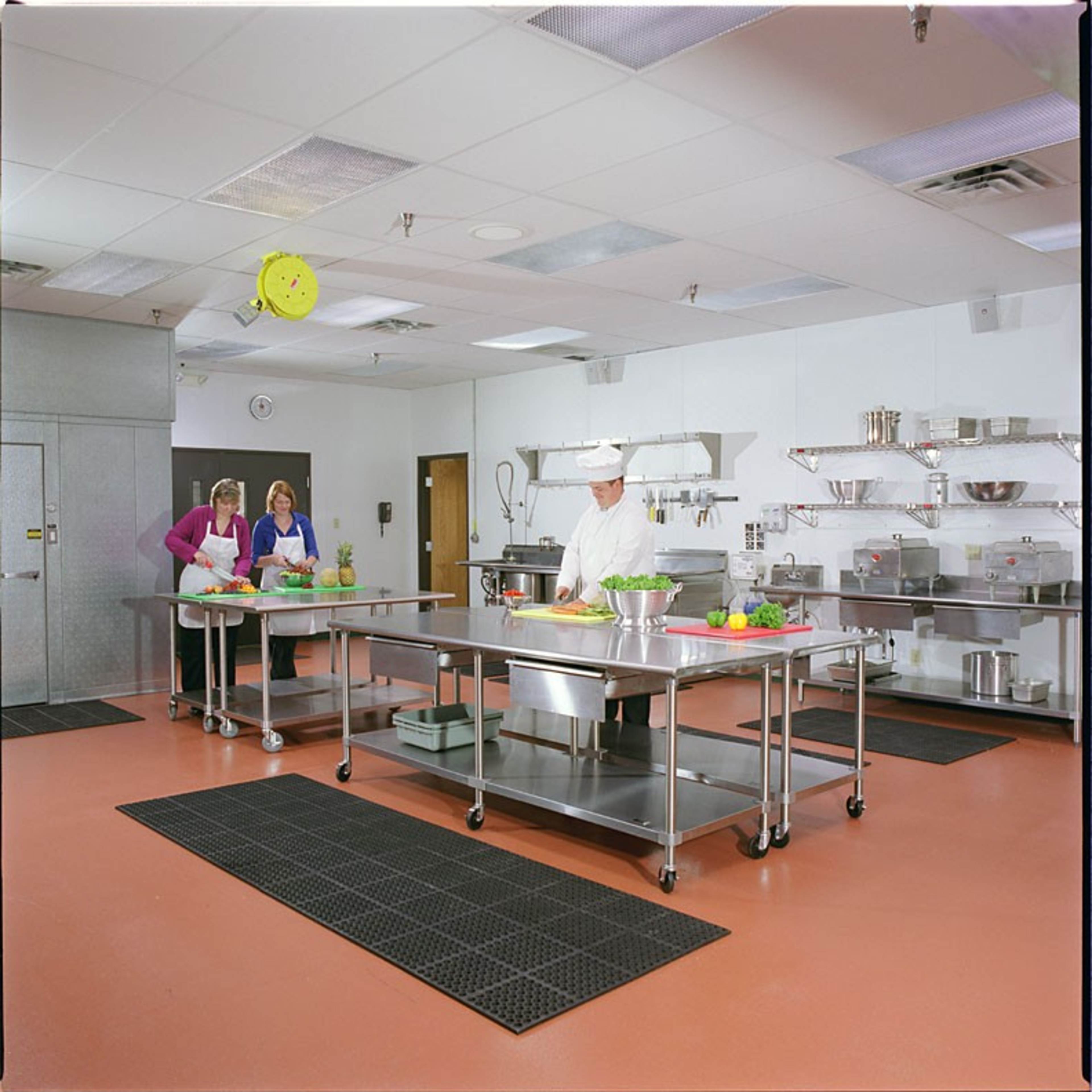 A clean, organized kitchen with stainless steel work tables, a chef in a white uniform, and two women preparing ingredients nearby.