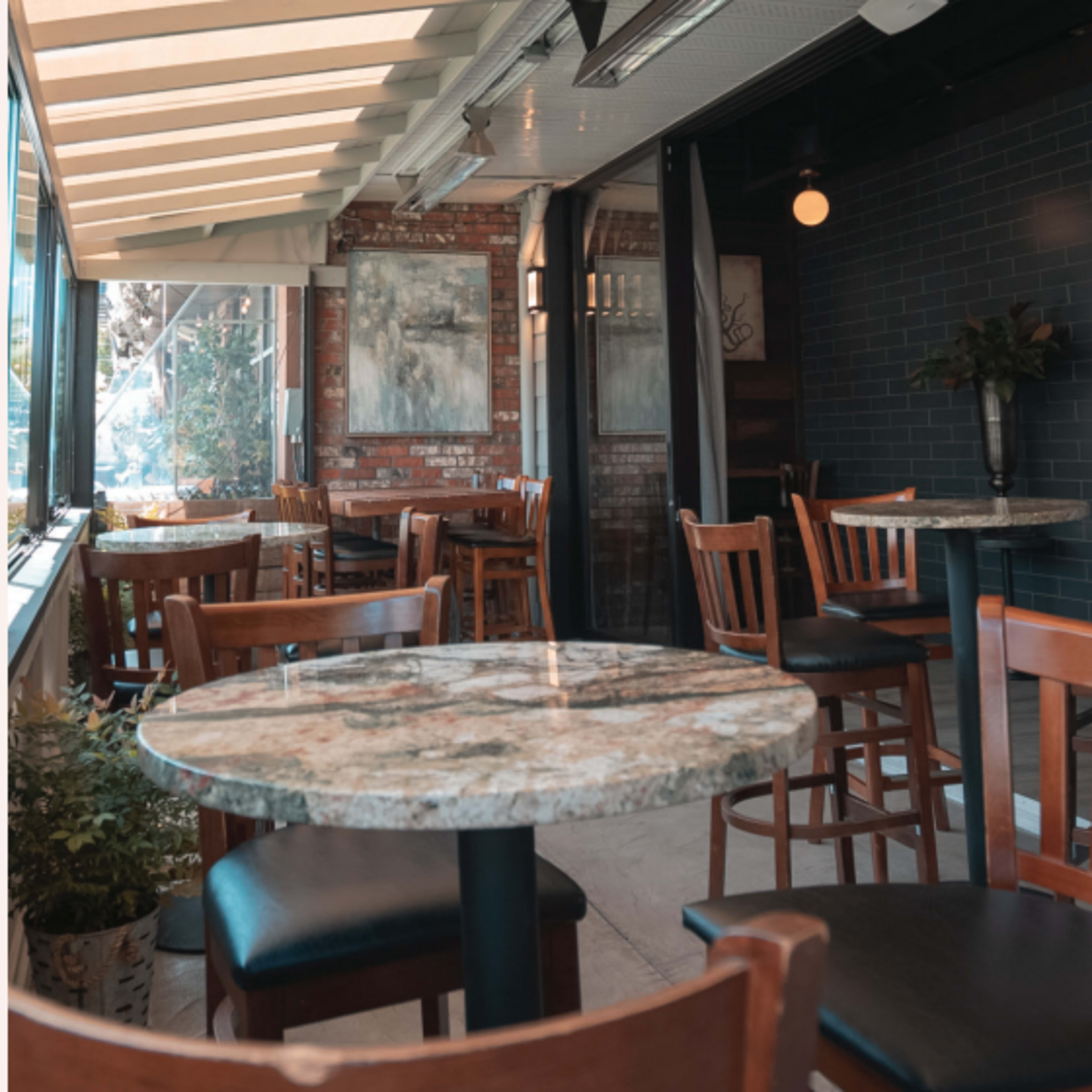A bright, inviting café with wooden tables and chairs arranged near large windows and exposed brick walls.