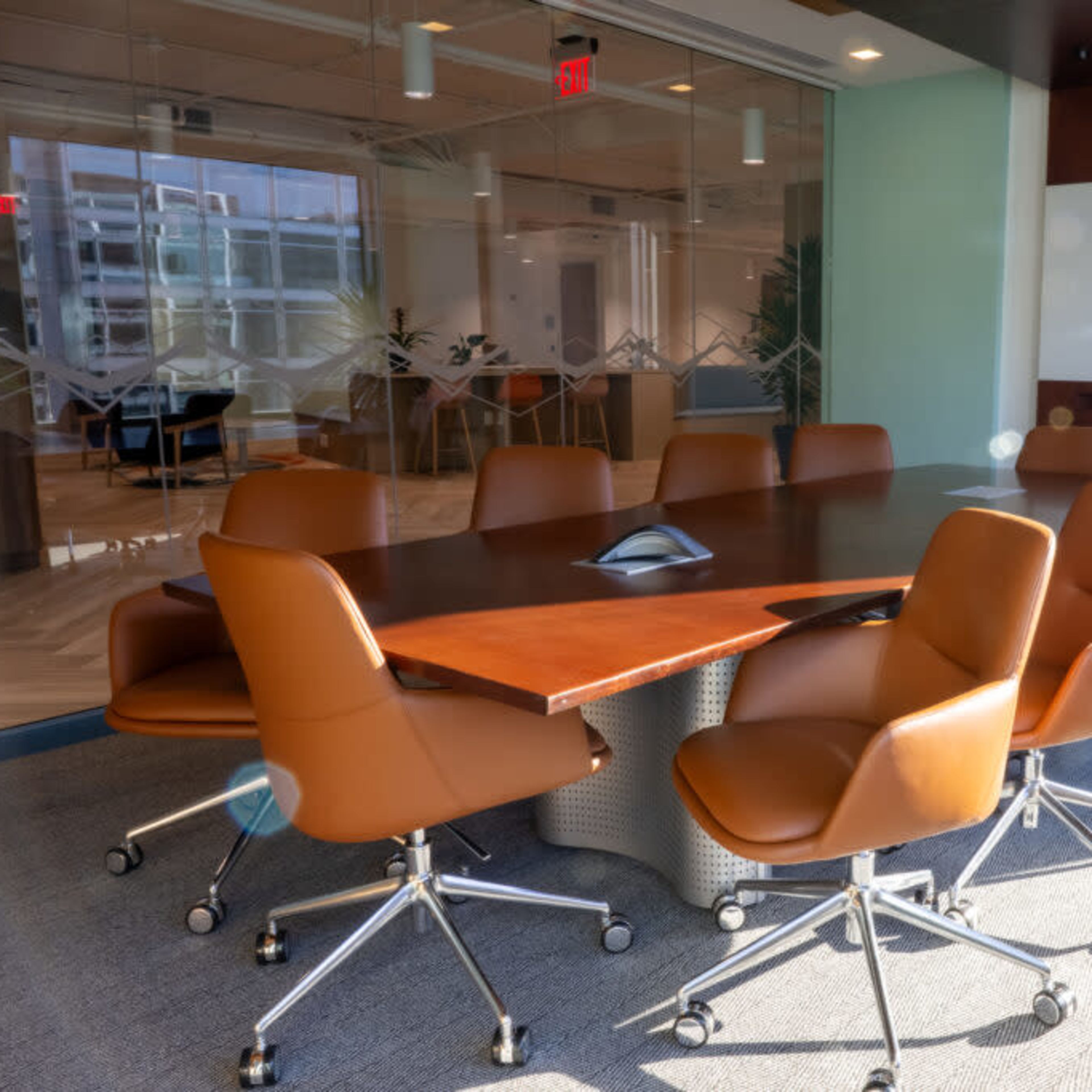 A modern conference room features a long wooden table surrounded by eight brown leather chairs, with a large window letting in natural light.