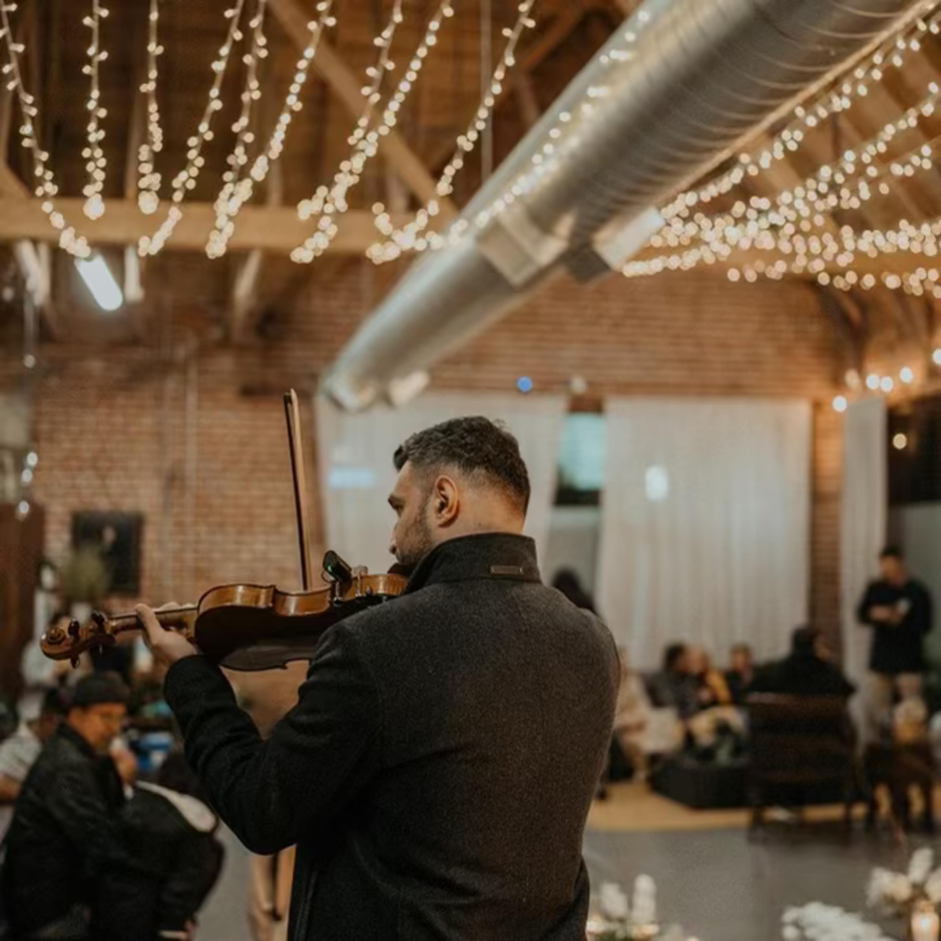 A musician plays the violin in a spacious venue decorated with string lights and flowers, while an audience sits in the background.