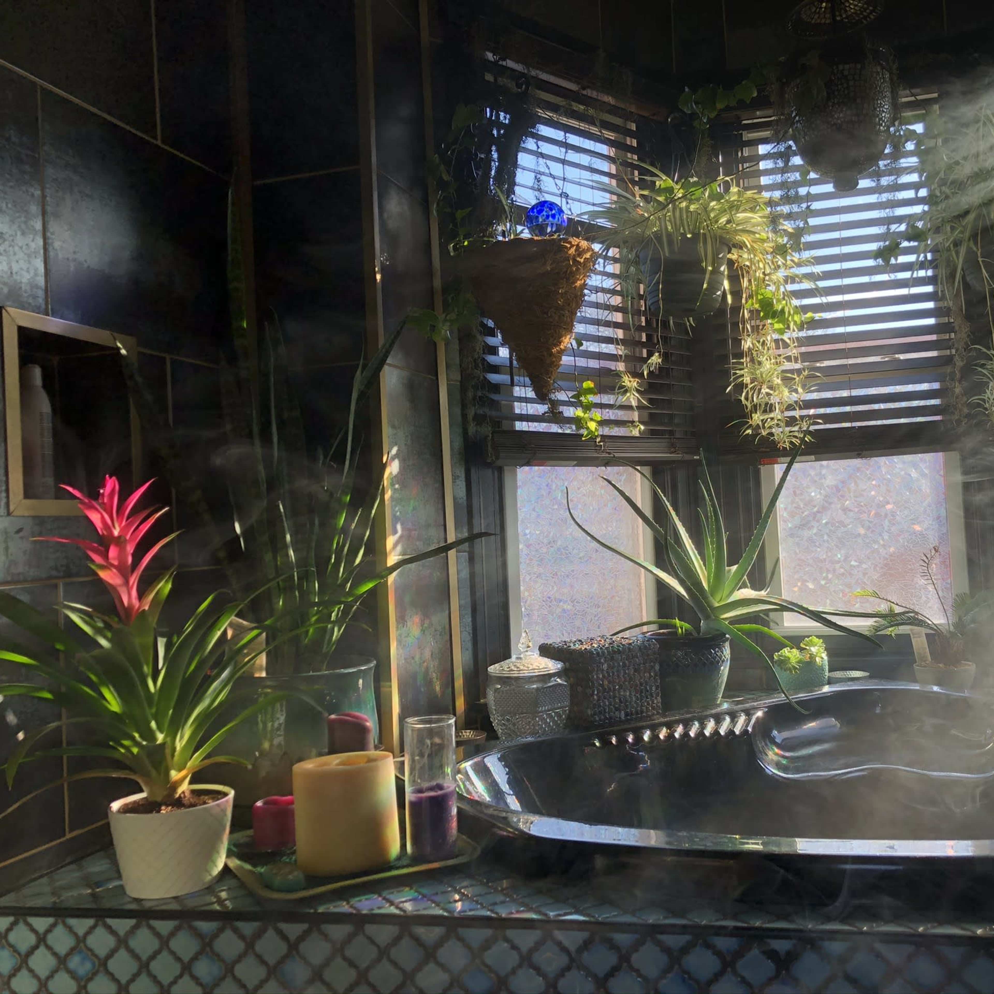A sunlit bathroom with a black tub surrounded by various plants and candles, with steam rising in the air.
