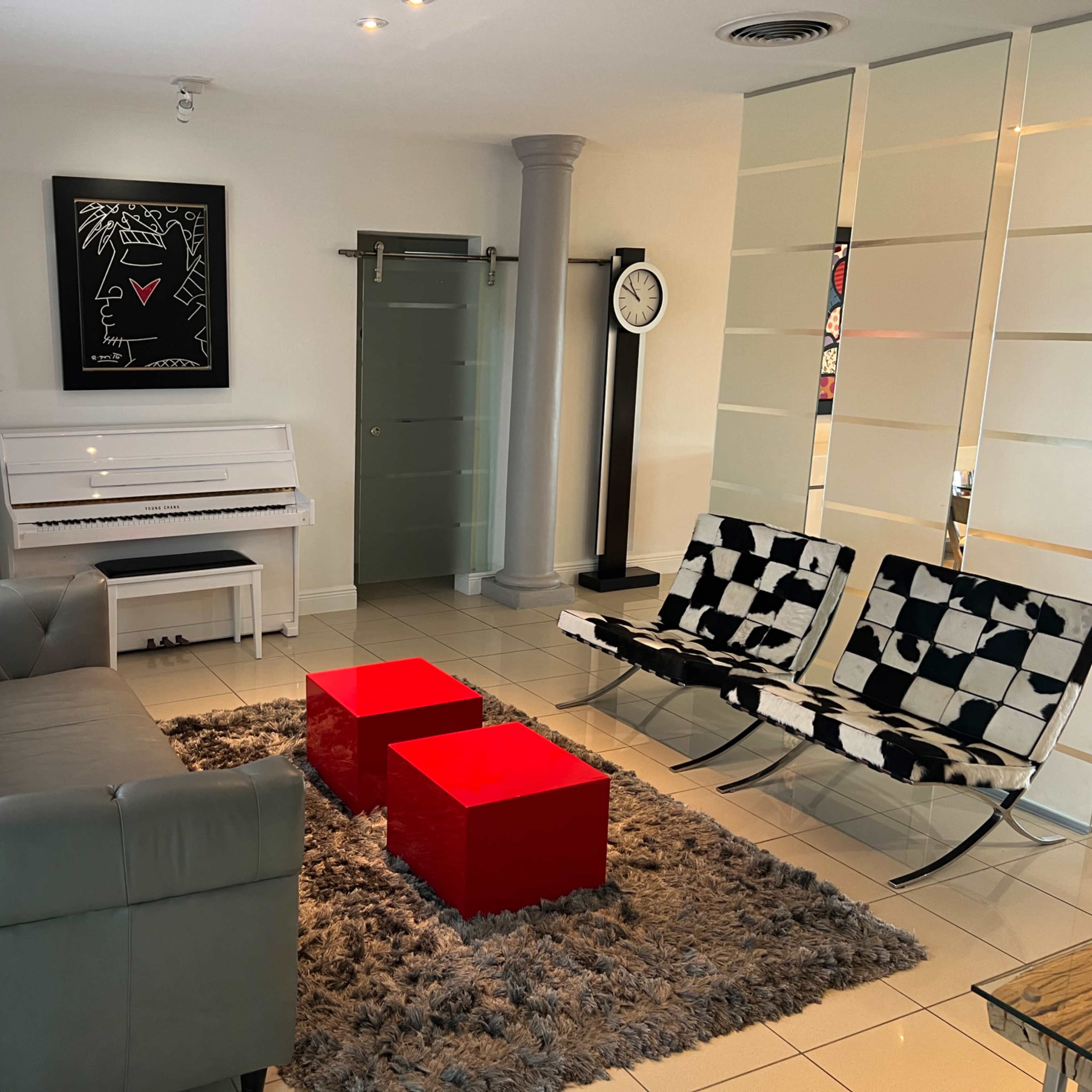 A modern living room with a white piano, two red square coffee tables, black and white patterned chairs, and a gray sectional sofa on a rug.