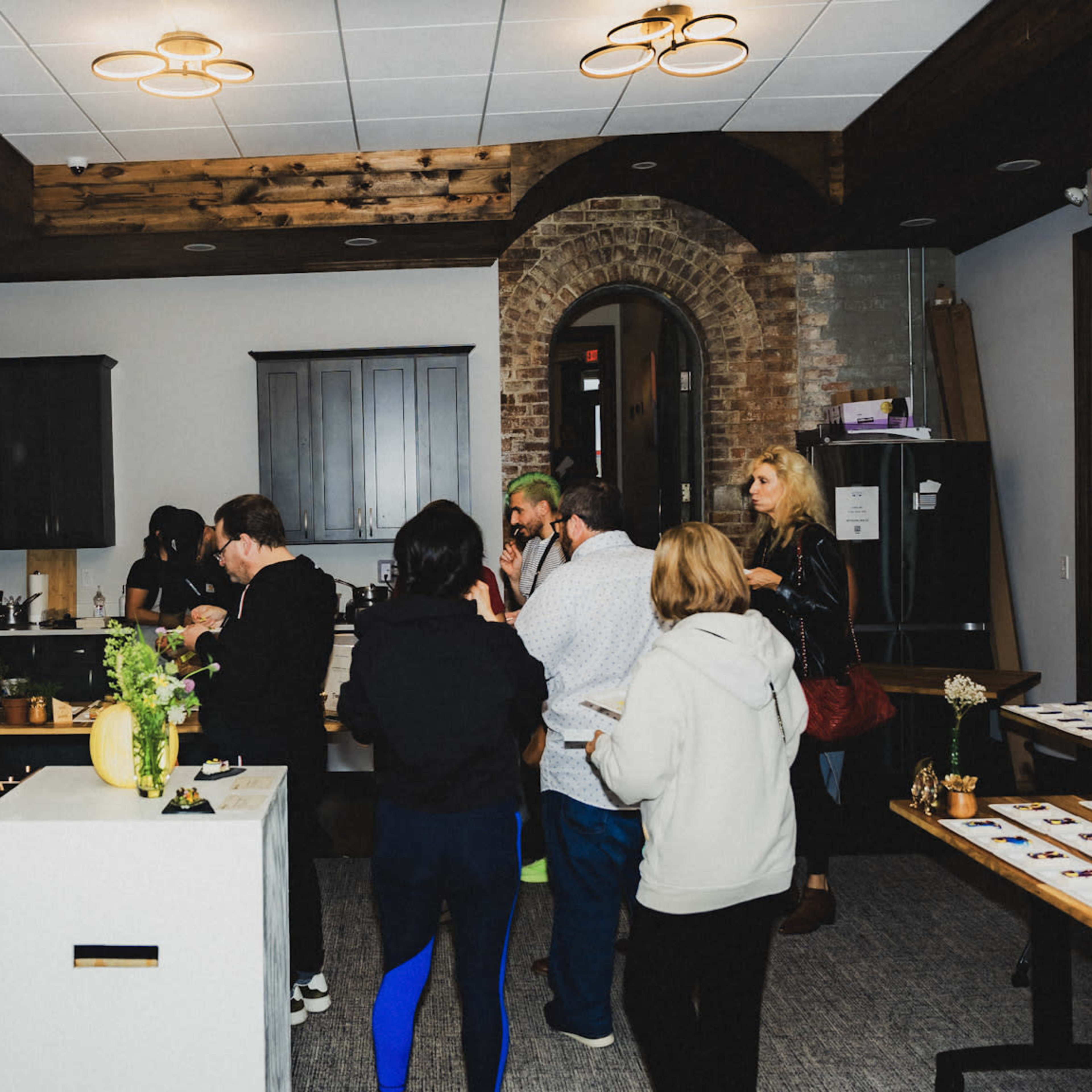 A group of people are gathered in a spacious room with dark wood accents, engaging in conversation and sampling food at a modern event.