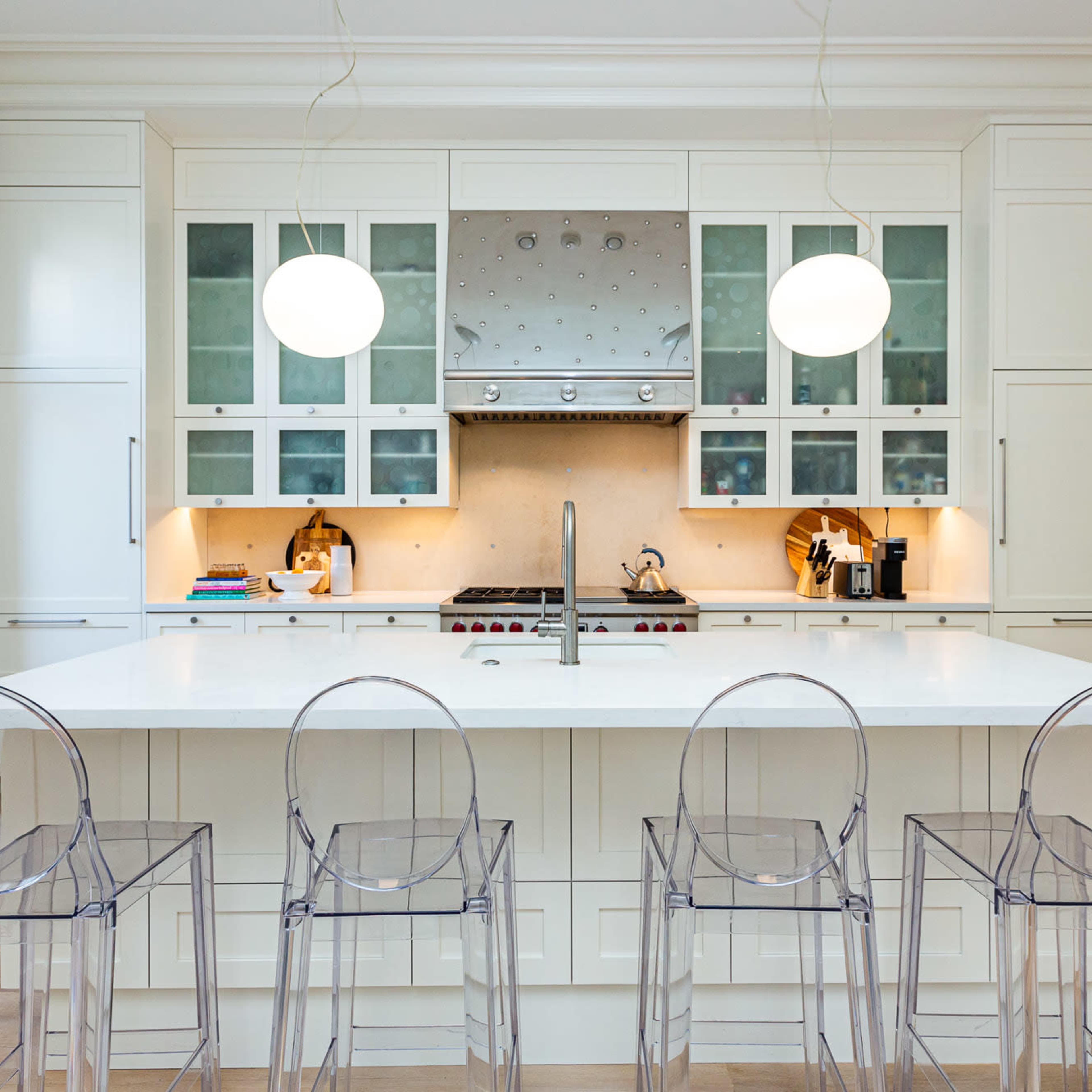 A modern kitchen features a large white island with four clear acrylic bar stools and a stainless steel range hood above a cooking area.