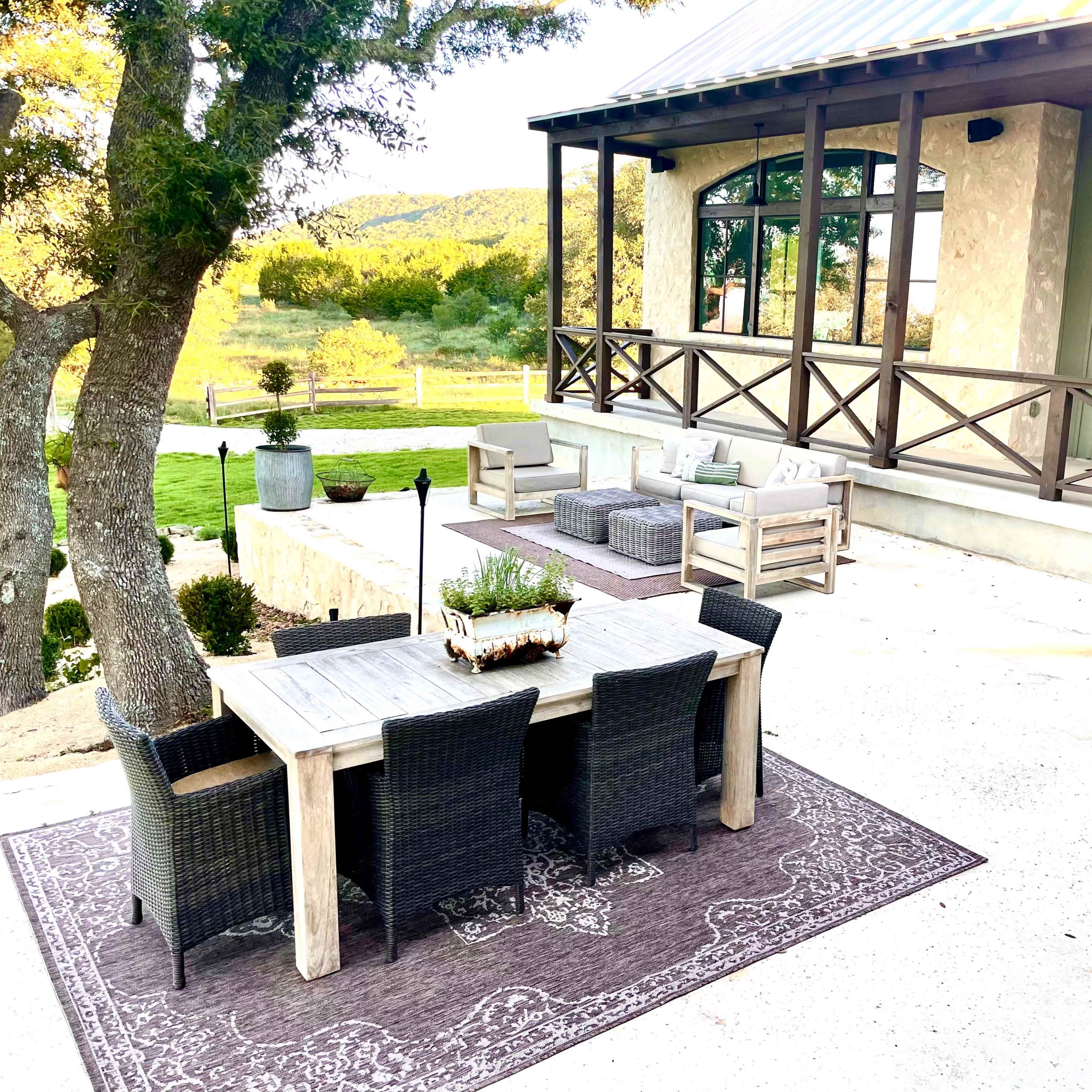 A rustic outdoor dining area features a wooden table surrounded by black wicker chairs on a concrete patio, with a backdrop of a house and greenery.