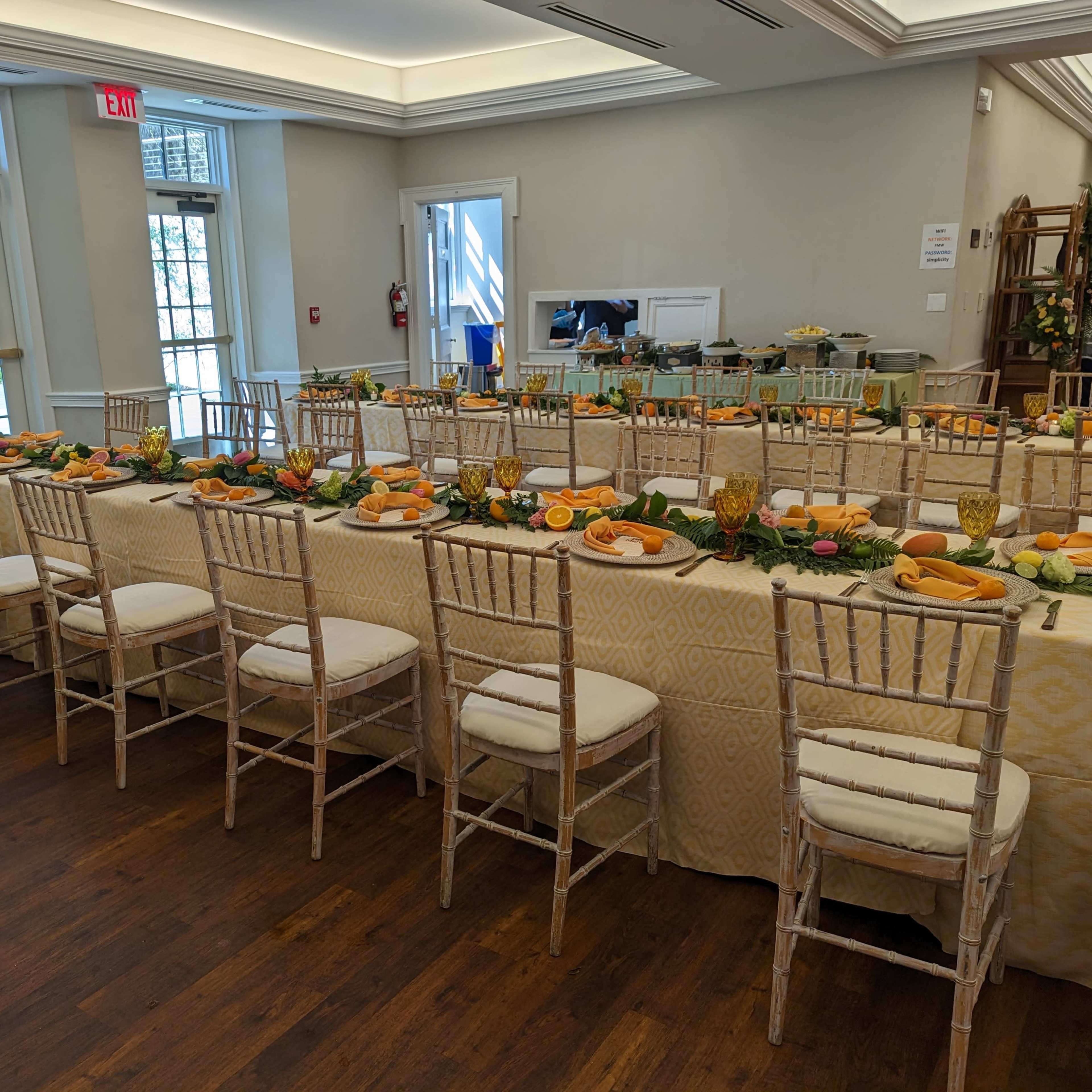 A dining setup features a long table adorned with orange fruits, greenery, and glassware in a well-lit room with large windows.