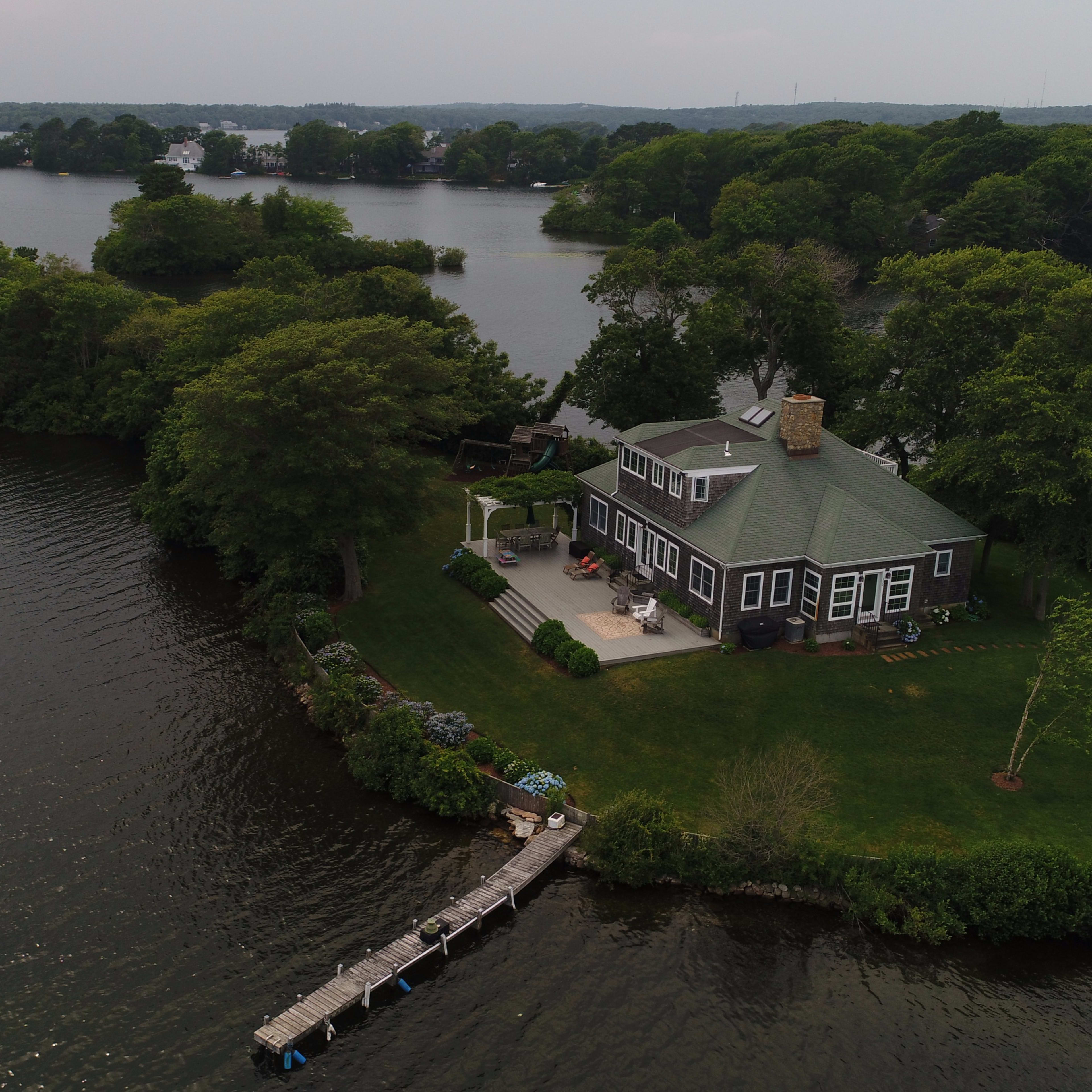 A large house with a green roof is situated on a small island surrounded by water, featuring a dock extending into the lake.