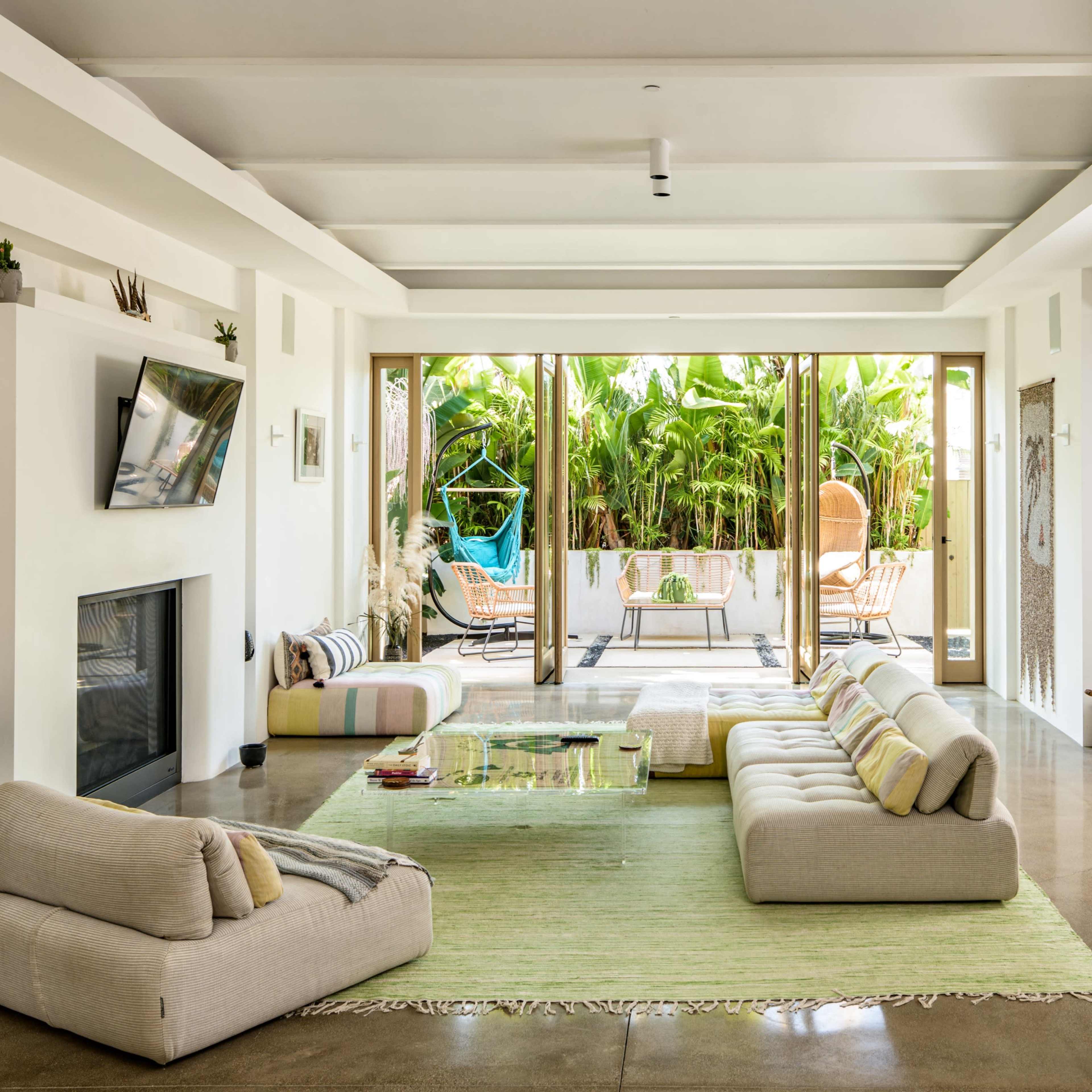 A spacious living room features a low seating area with a glass table, large windows opening to a patio with lush greenery.