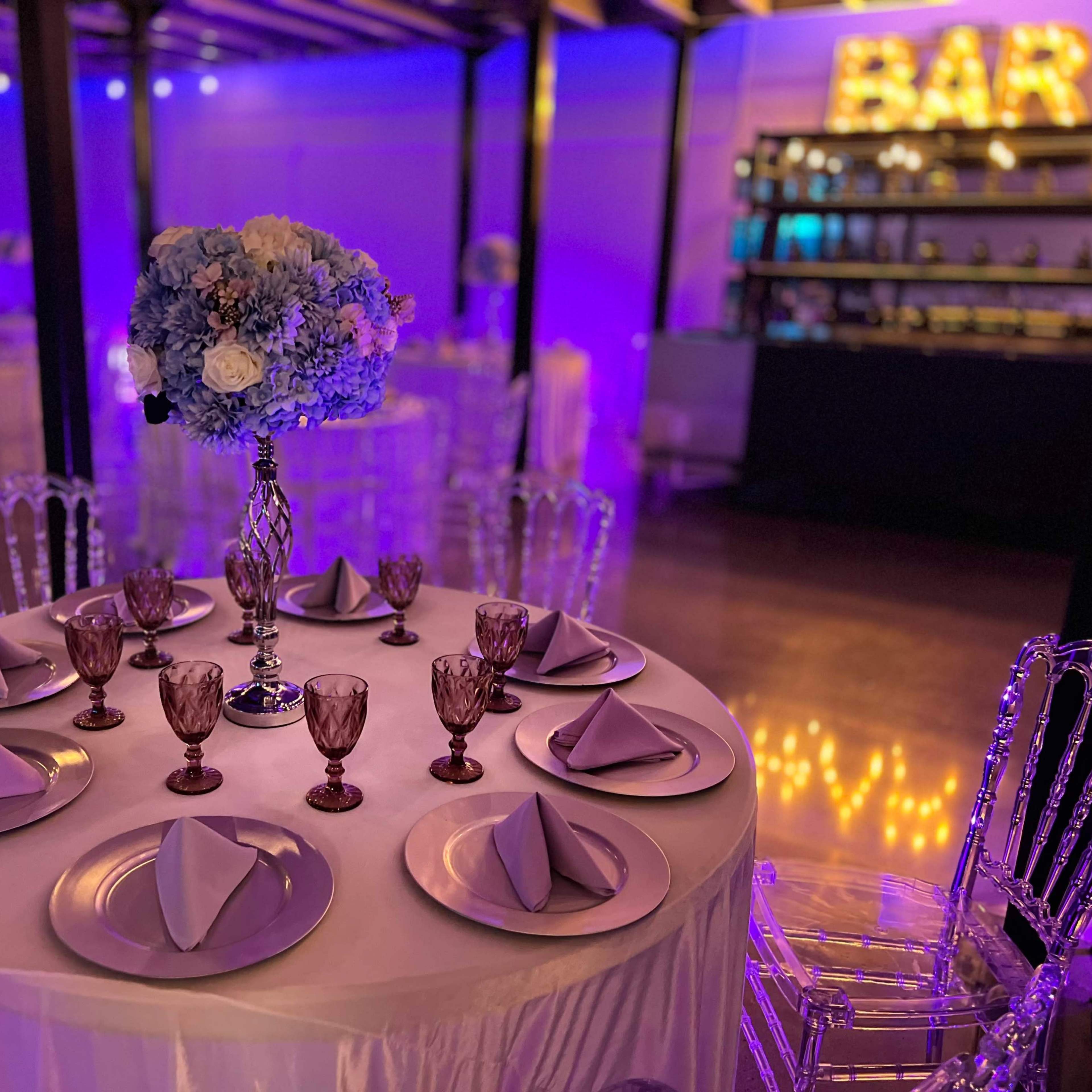 A round table is elegantly set with folded napkins and glasses, surrounded by transparent chairs in a softly lit venue featuring a bar in the background.