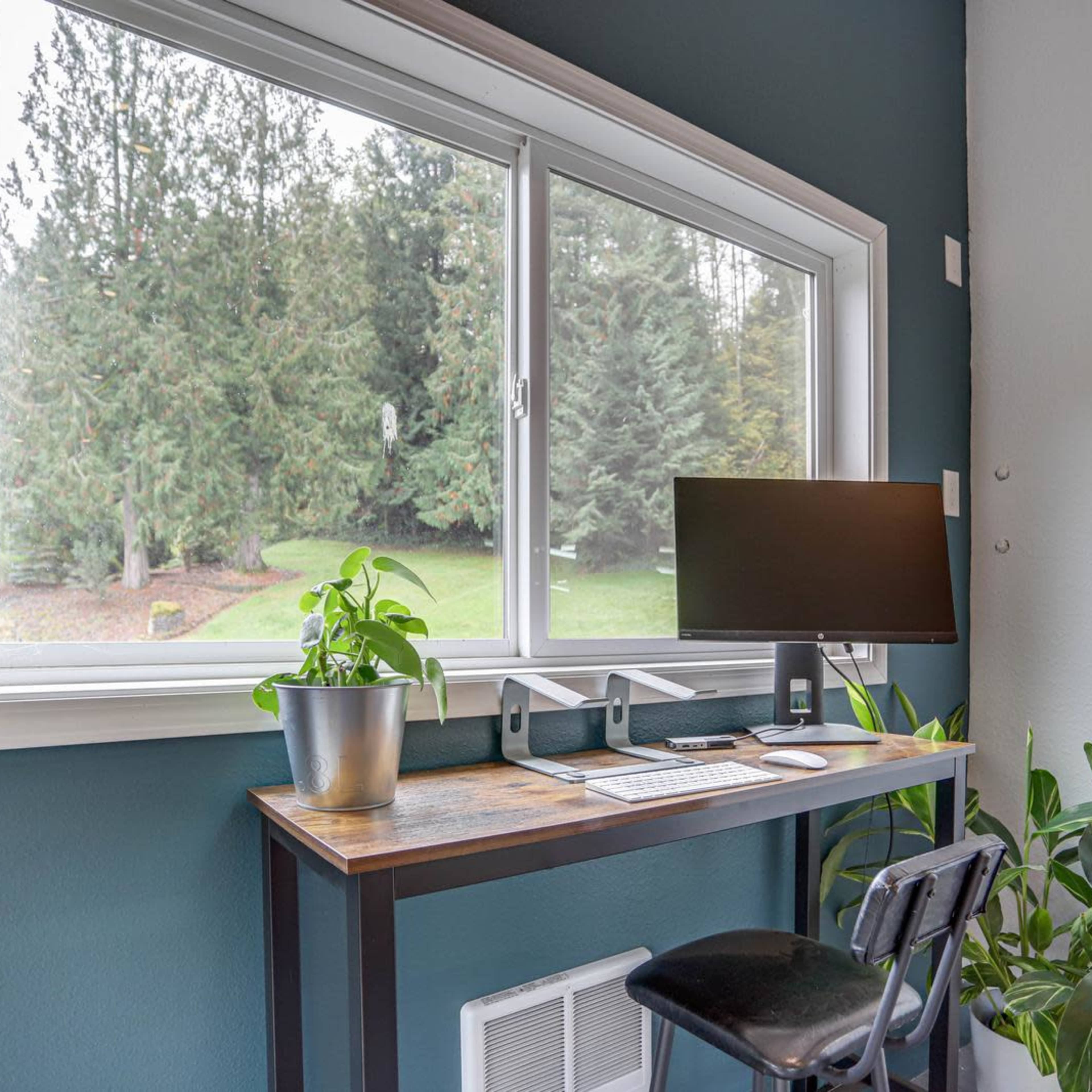 A modern workspace with a computer monitor, keyboard, and a potted plant, situated by a large window overlooking a green landscape.