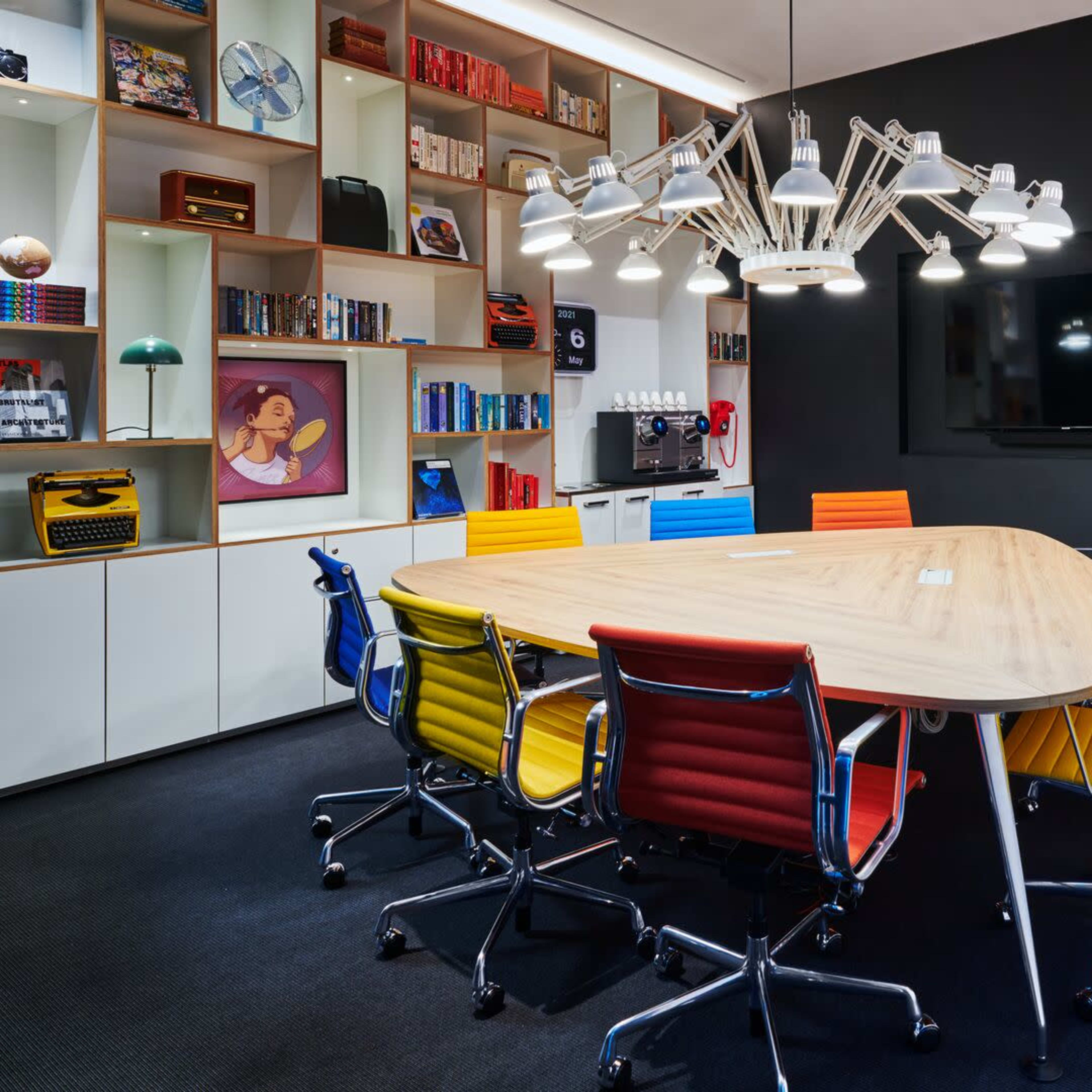 A modern conference room features an oval wooden table surrounded by colorful chairs and a wall-mounted TV, with bookshelves and decorative items in the background.