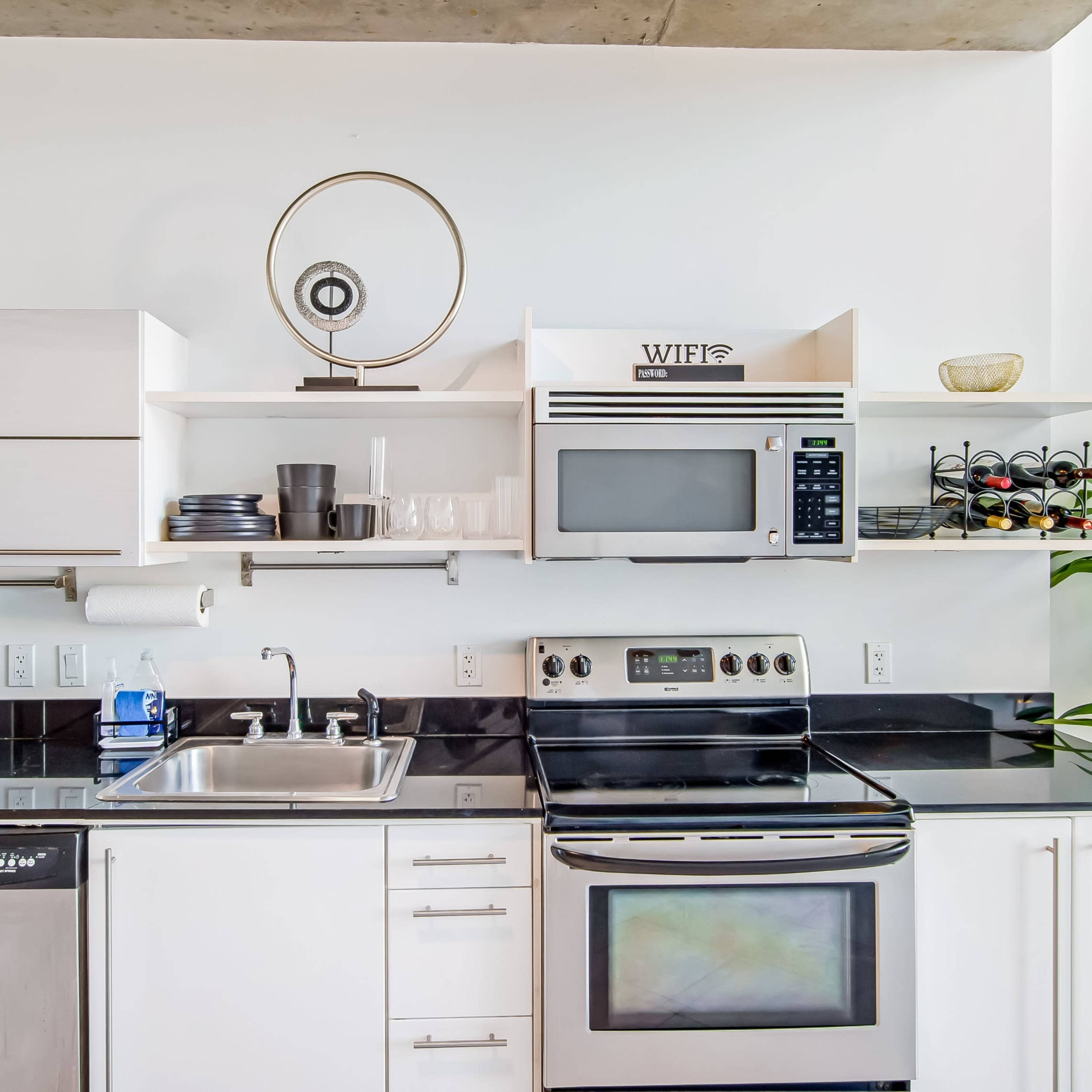 A modern kitchen features black countertops, stainless steel appliances, a wall-mounted microwave, and open shelving with dishware and a decorative plant.