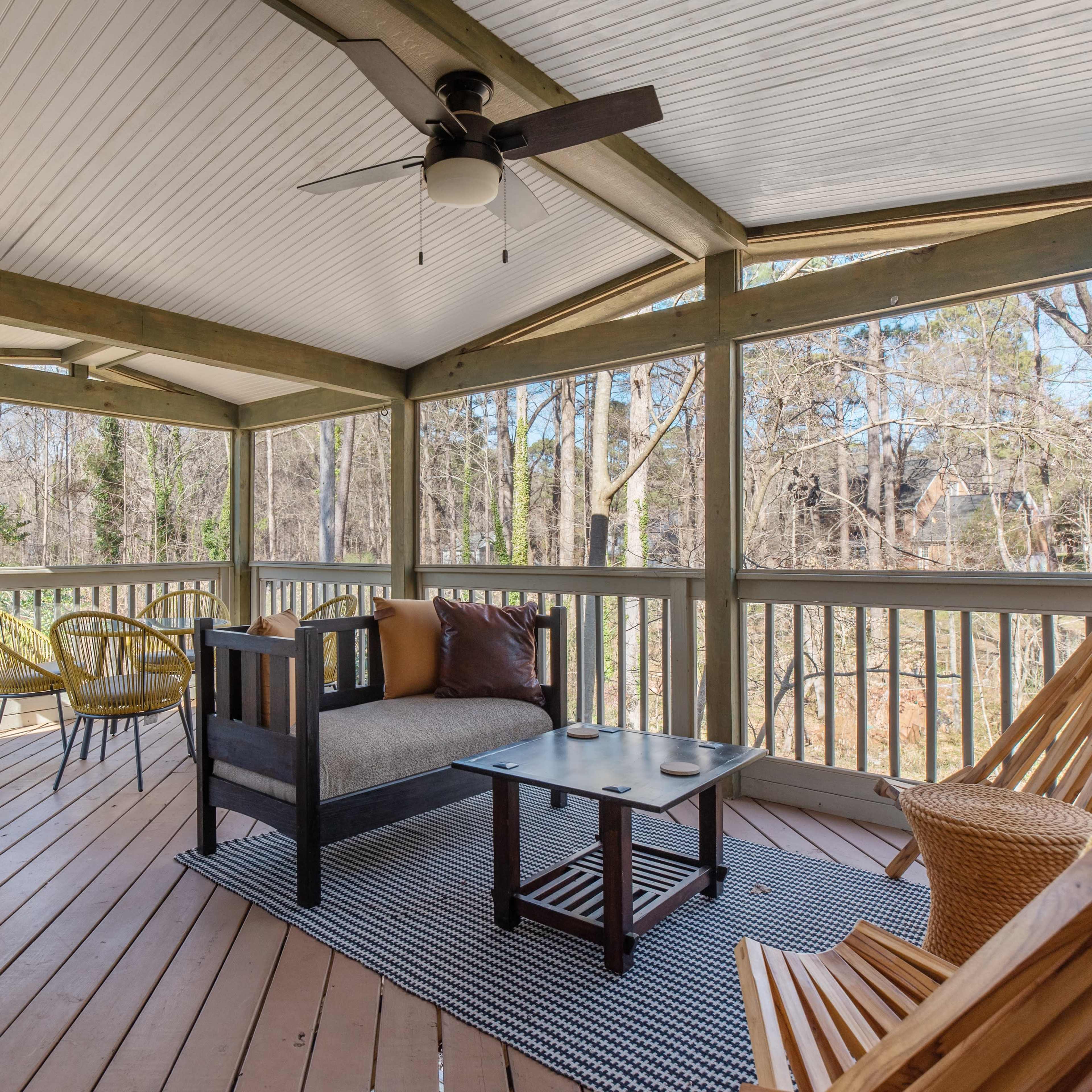 A screened porch with seating arrangements, including a sofa, coffee table, and various chairs, surrounded by trees.