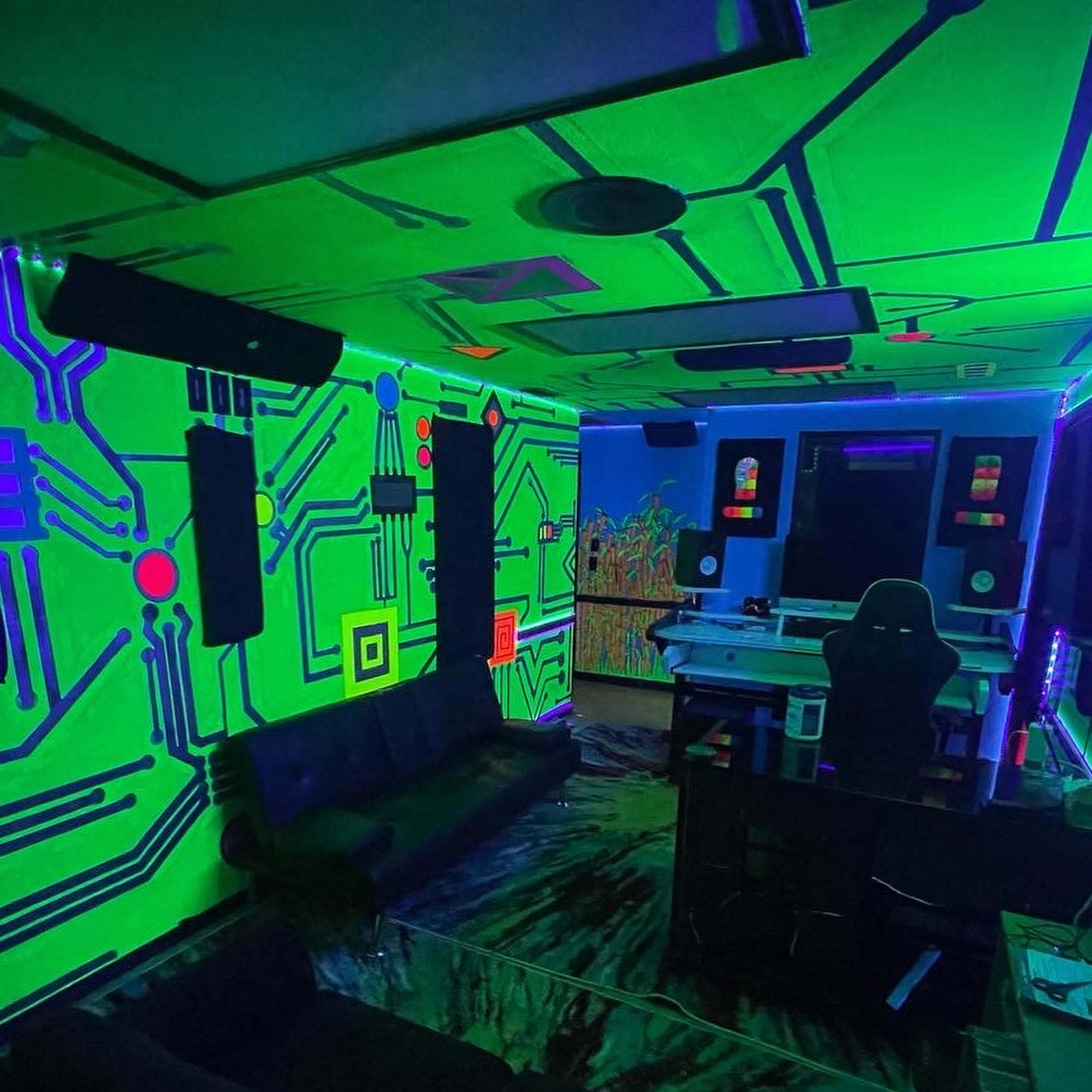 The room features neon circuit board designs on the walls, with black couches and a desk illuminated by colorful lighting.