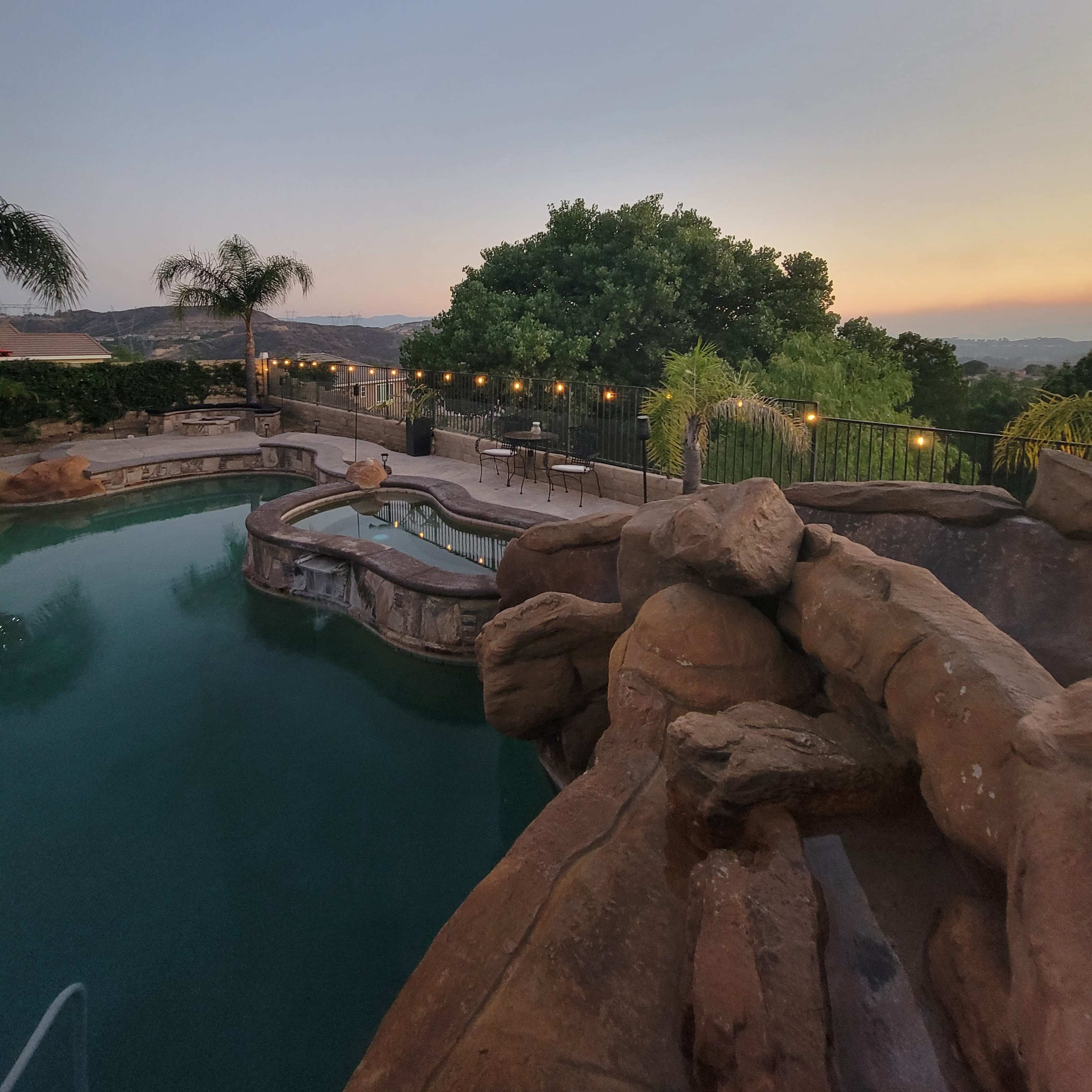 A landscaped backyard features a pool with a rocky slide, surrounded by palm trees and illuminated by string lights at dusk.