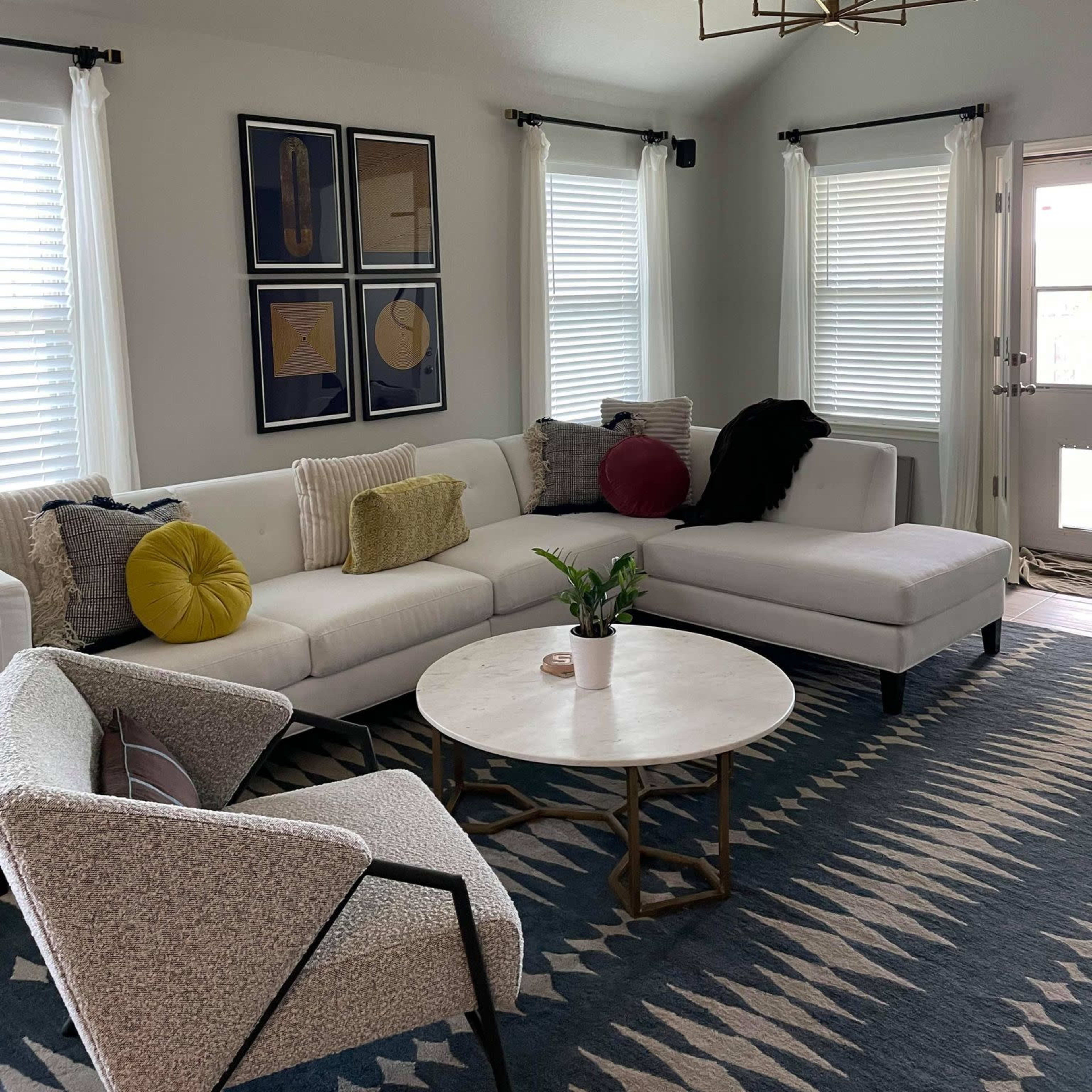 A modern living room features a white sectional sofa, a round marble coffee table, and a patterned rug, with artwork on the walls and large windows letting in natural light.
