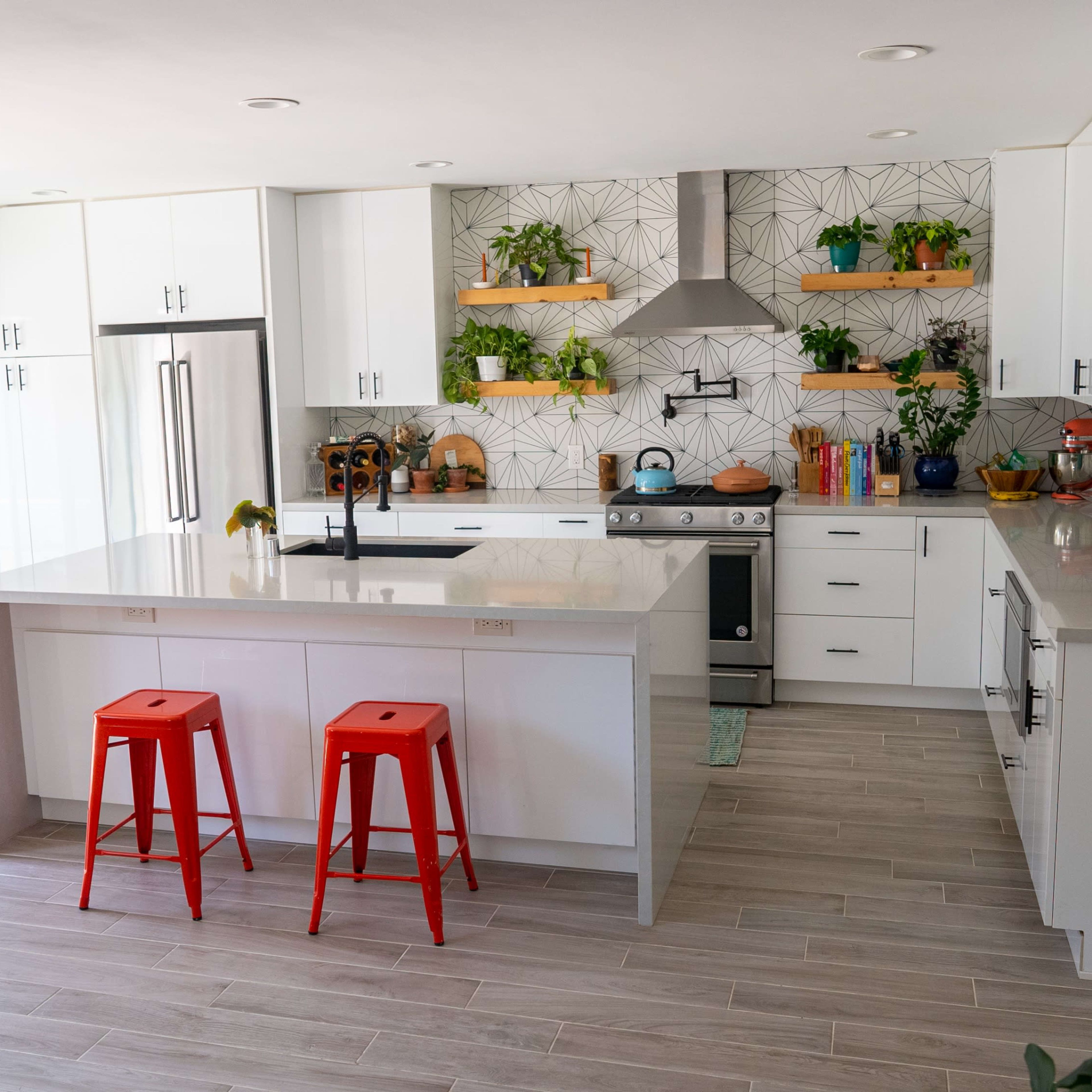 A modern kitchen with white cabinets, a central island with red stools, and plants arranged on shelves and countertops.