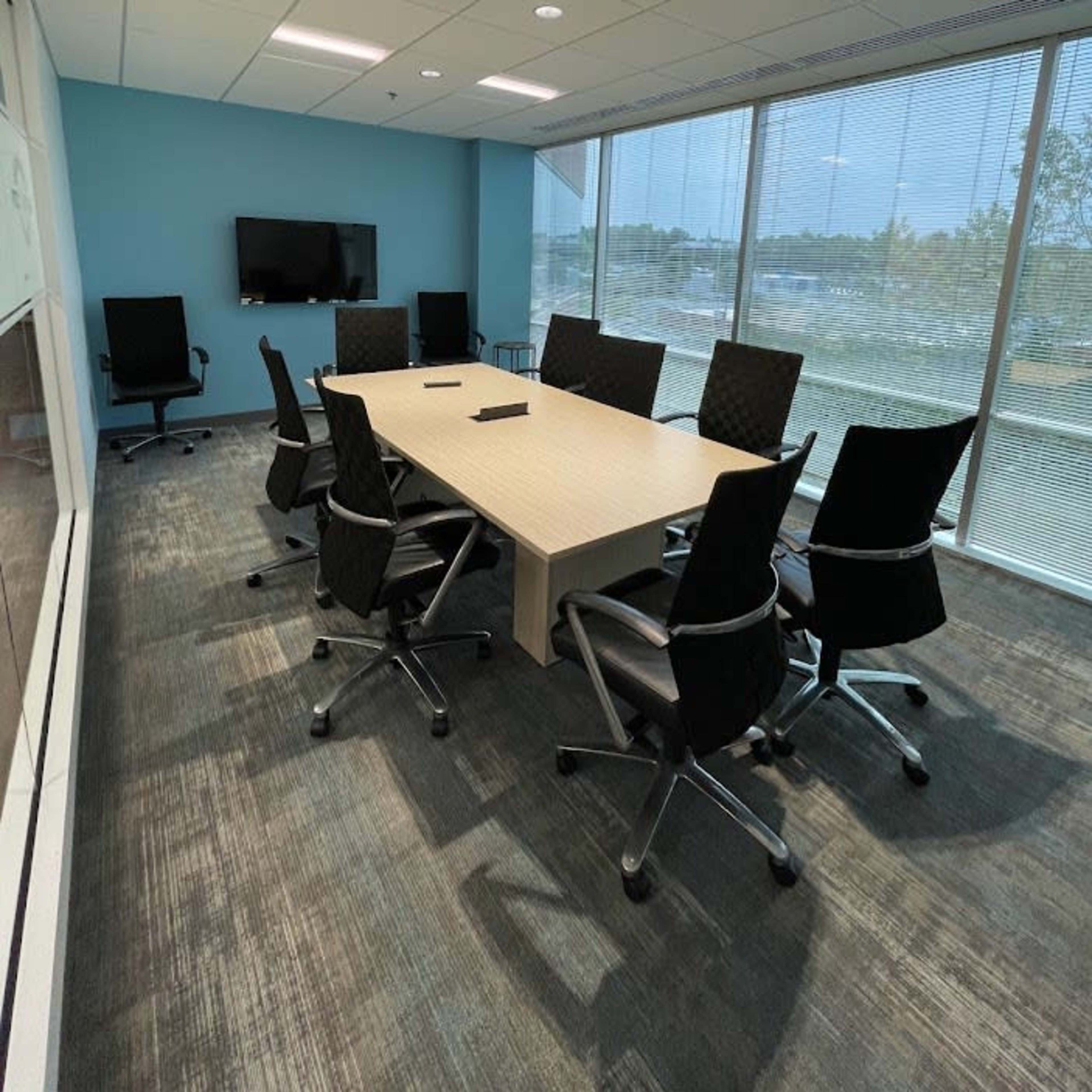 A conference room features a large table surrounded by ergonomic chairs, with a television mounted on the wall and windows letting in natural light.