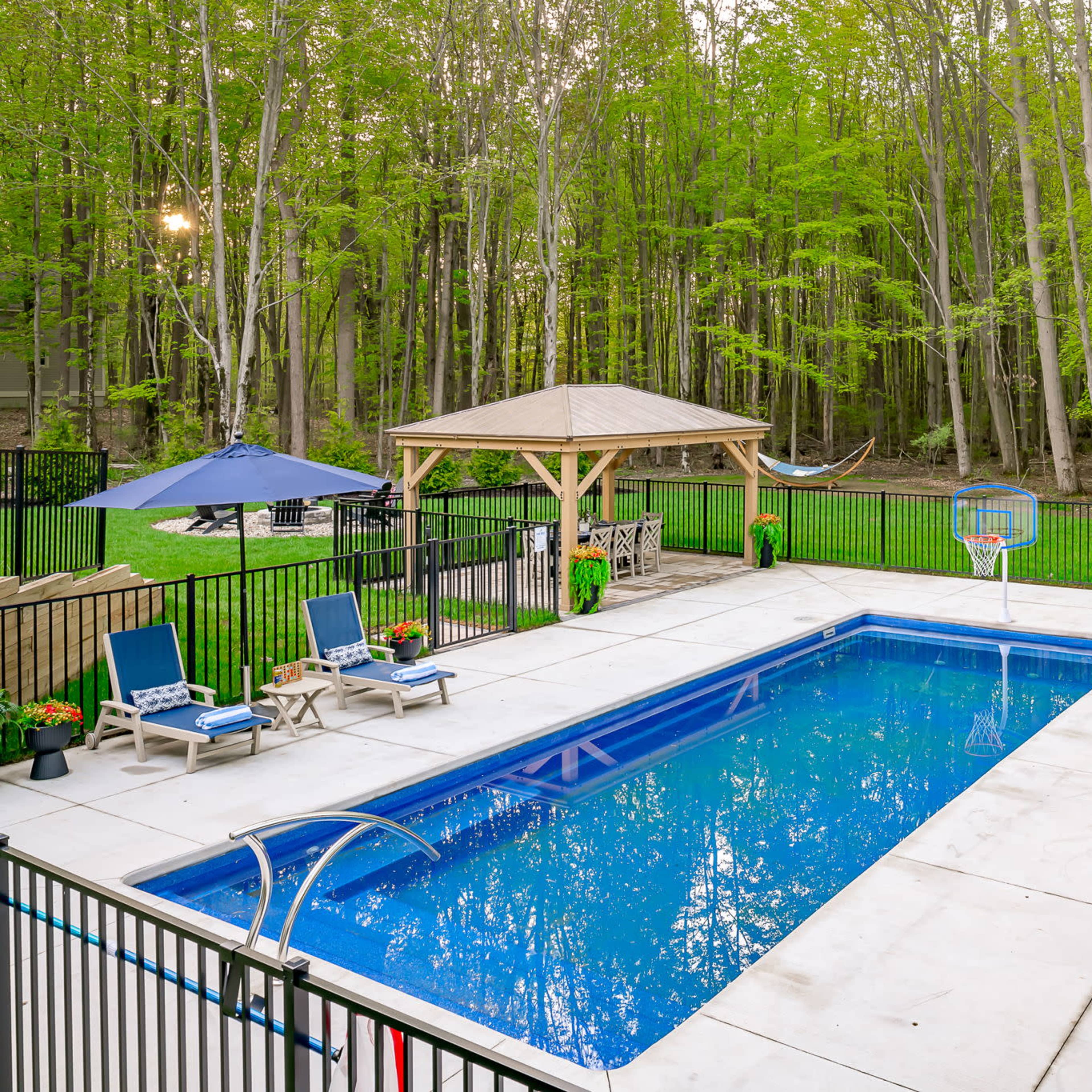 A rectangular swimming pool surrounded by lounge chairs and umbrellas, set in a green, wooded backyard with a gazebo and a hammock.